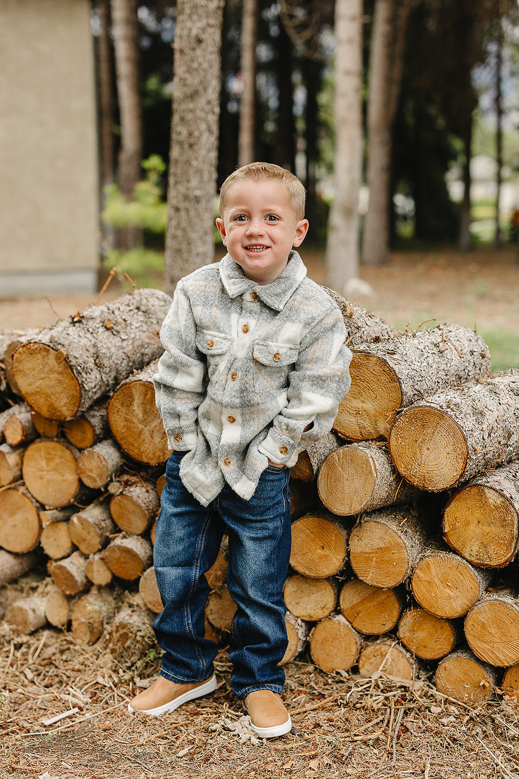 A boy wearing a grey plaid kids shacket, standing by a stack of logs outdoors.