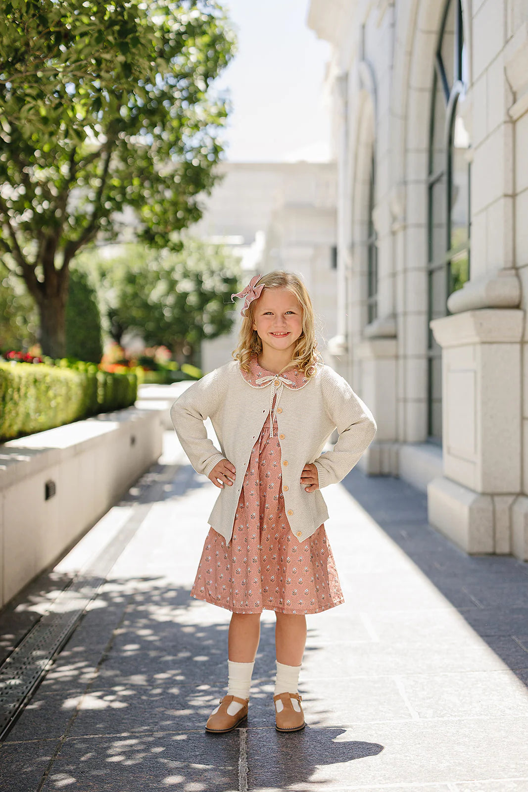 Little girl wearing an oatmeal knit button-down cardigan kids over a pink dress in a sunny outdoor setting.