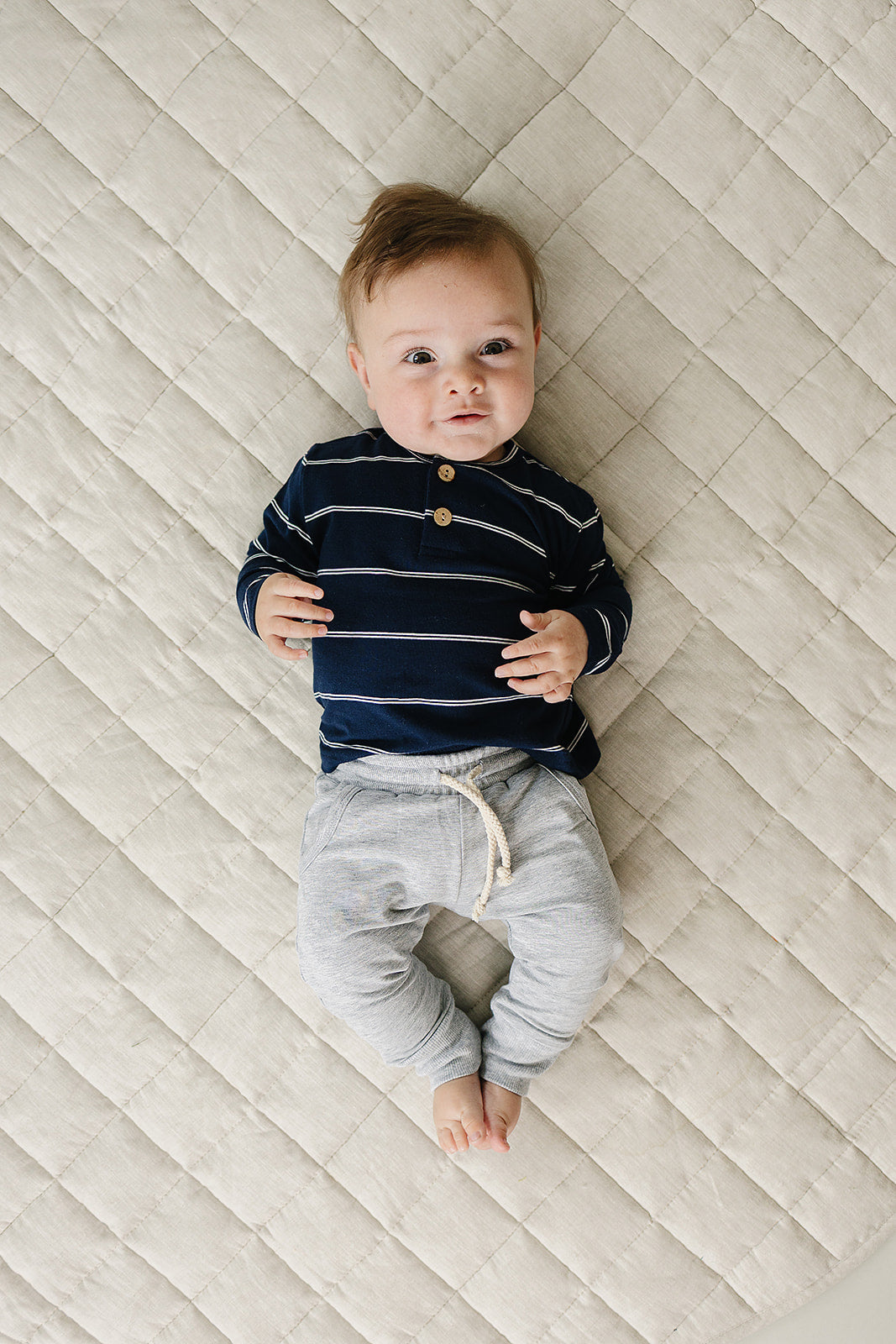 Cute baby wearing a navy stripe long sleeve tee for kids, lying on a quilted surface.