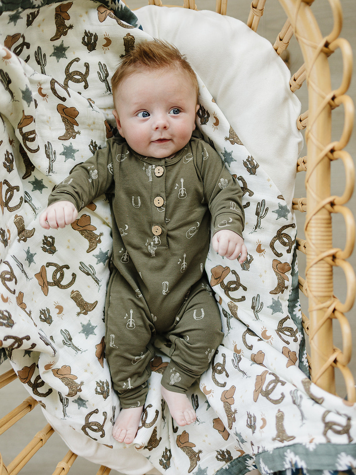 Baby in a cozy, olive green Western button romper surrounded by a playful printed blanket featuring cowboy themes.