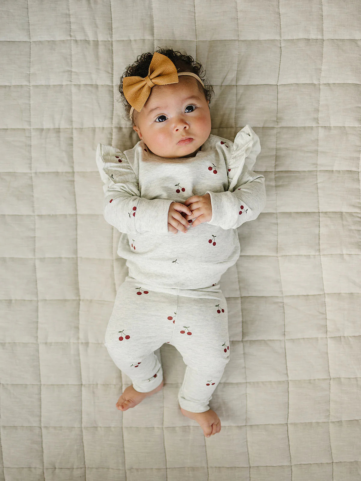 Baby girl in cherry ruffle French terry set with ruffle sleeves and cherry-print leggings, lounging on a quilted surface.