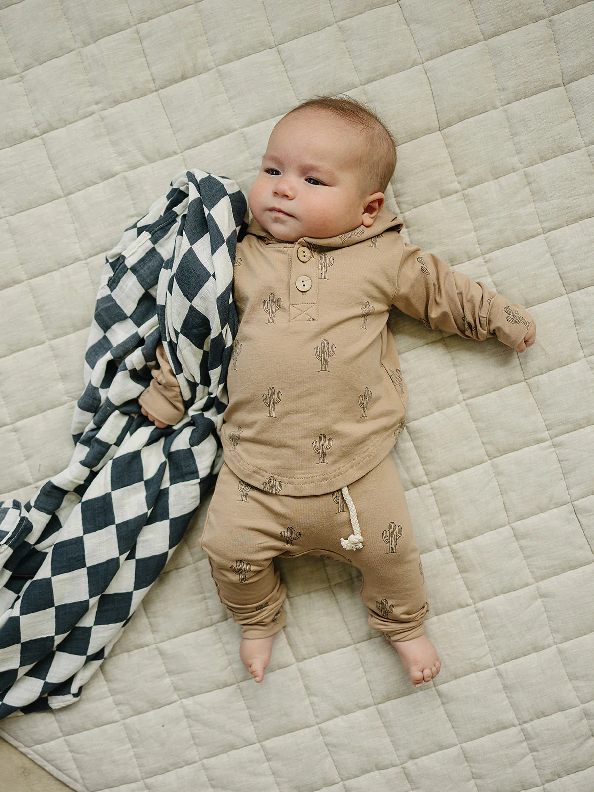 Baby in cactus hooded set kids, wearing a beige top and pants, with a blanket on a quilted surface.
