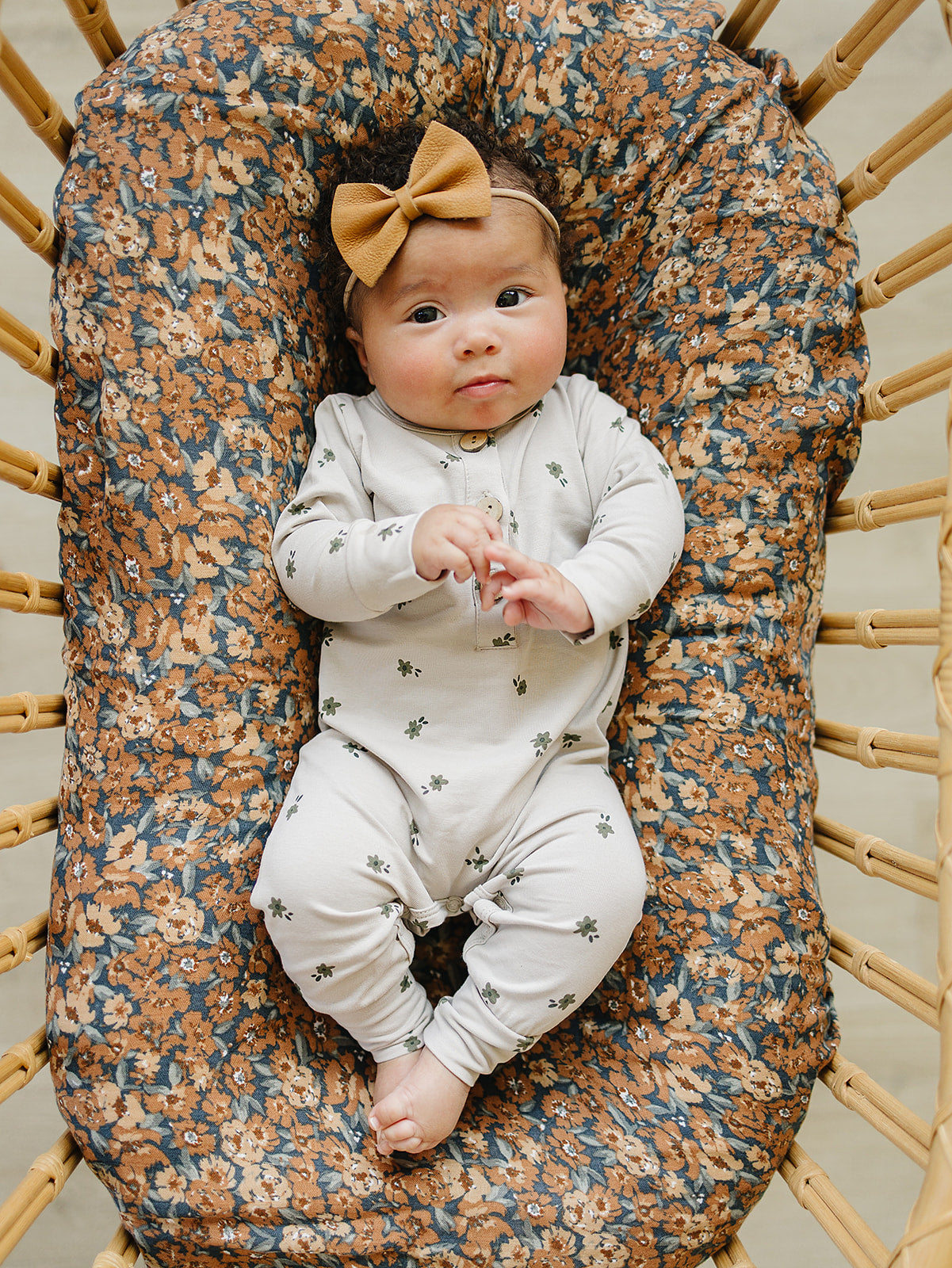 Baby girl in a floral button romper with long sleeves and a bow, lying in a cozy crib with floral bedding.