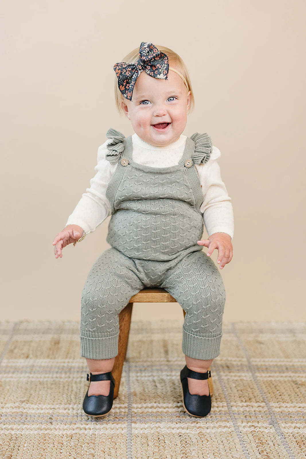 Cute baby girl smiling in green ruffle knit overalls, sitting on a wooden stool with a bow in her hair.