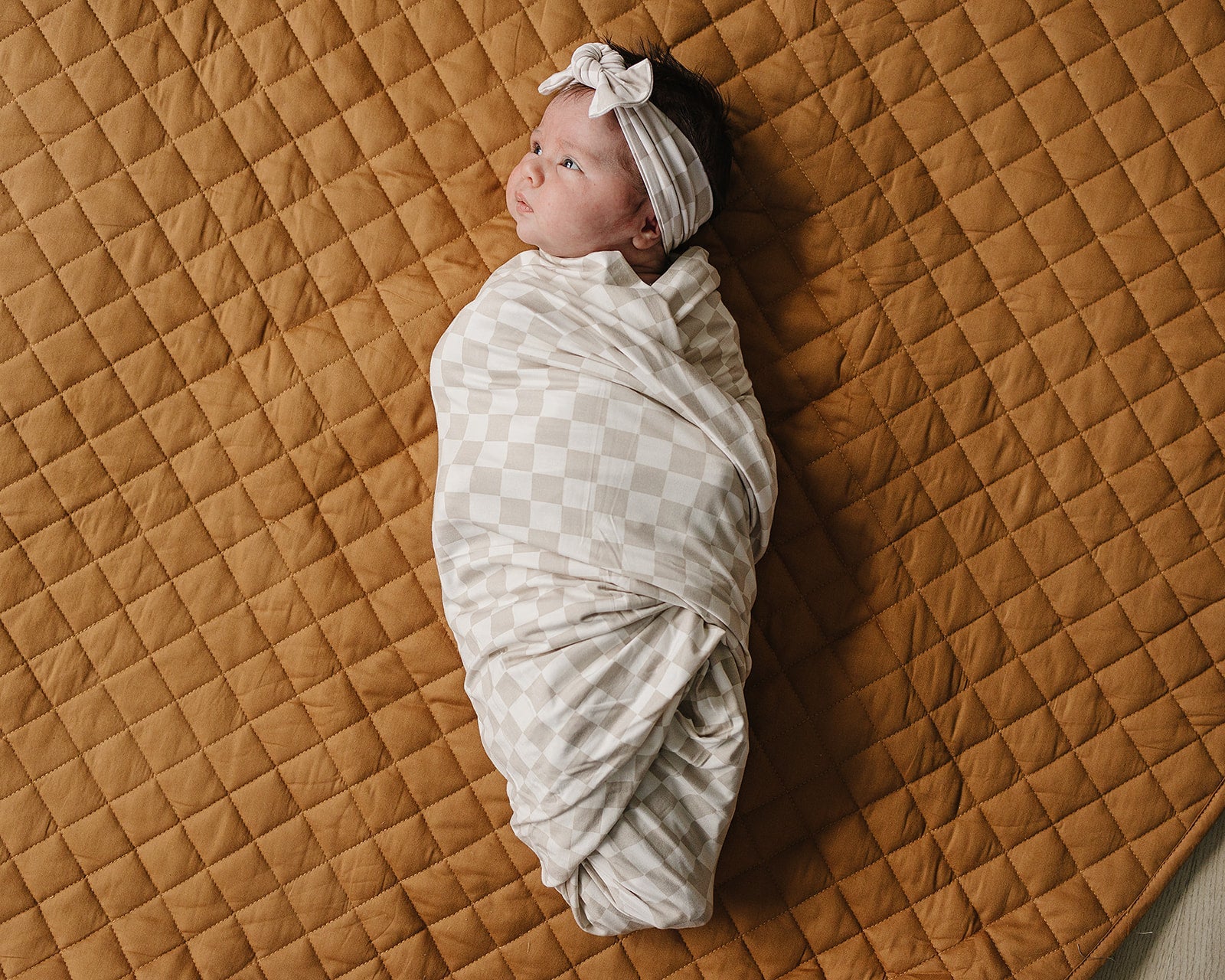 Bamboo Stretch Swaddle Taupe Checkered wrapped around a baby with a headband, resting on a cozy quilted blanket.