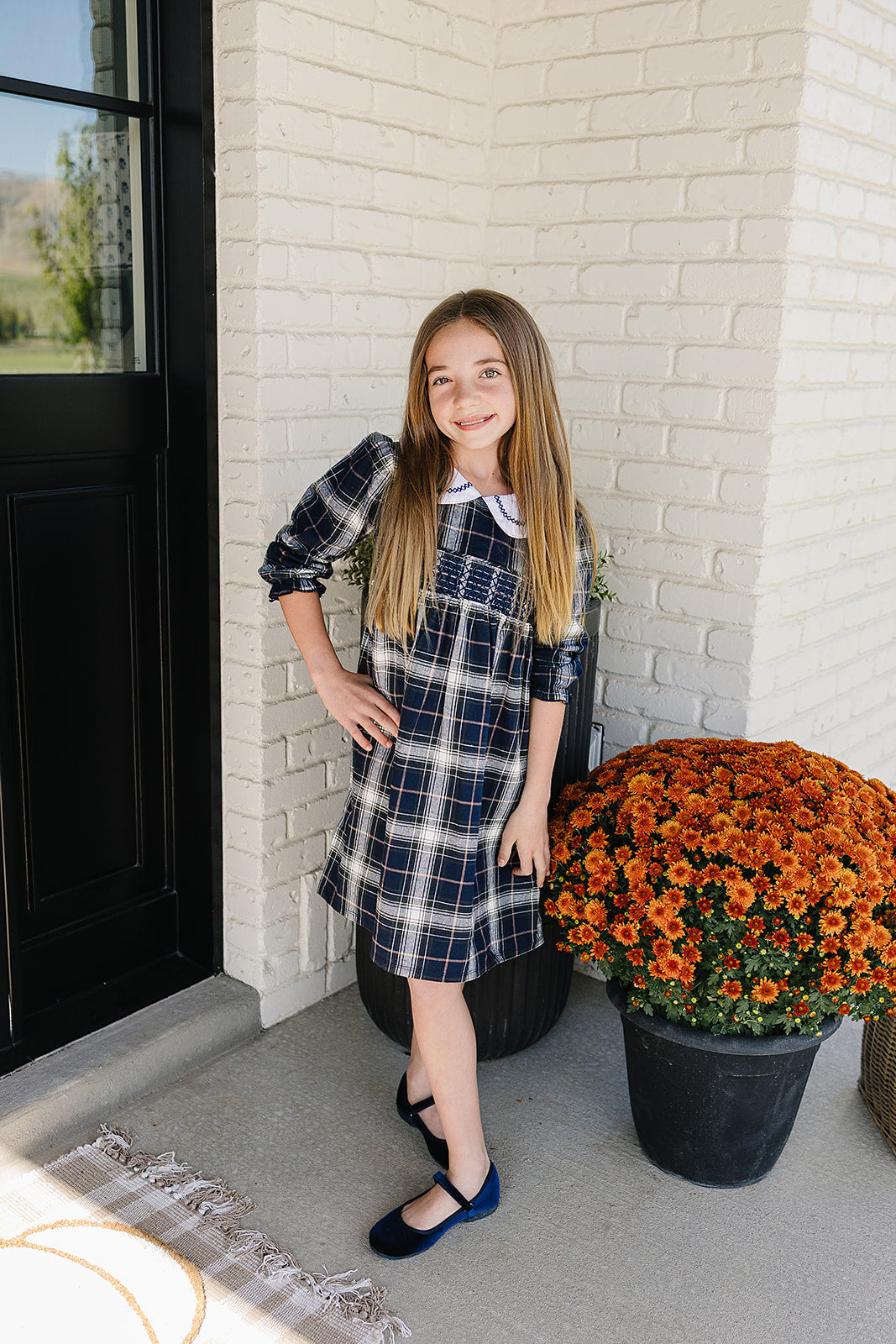 Girl wearing a navy plaid smocked dress with Peter Pan collar standing beside orange mums, showcasing a stylish girls plaid dress.