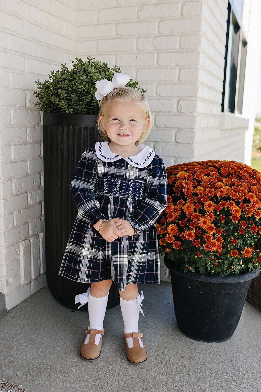 Cute girl wearing a Navy Plaid Smocked Dress with Peter Pan Collar, playing near orange flowers.