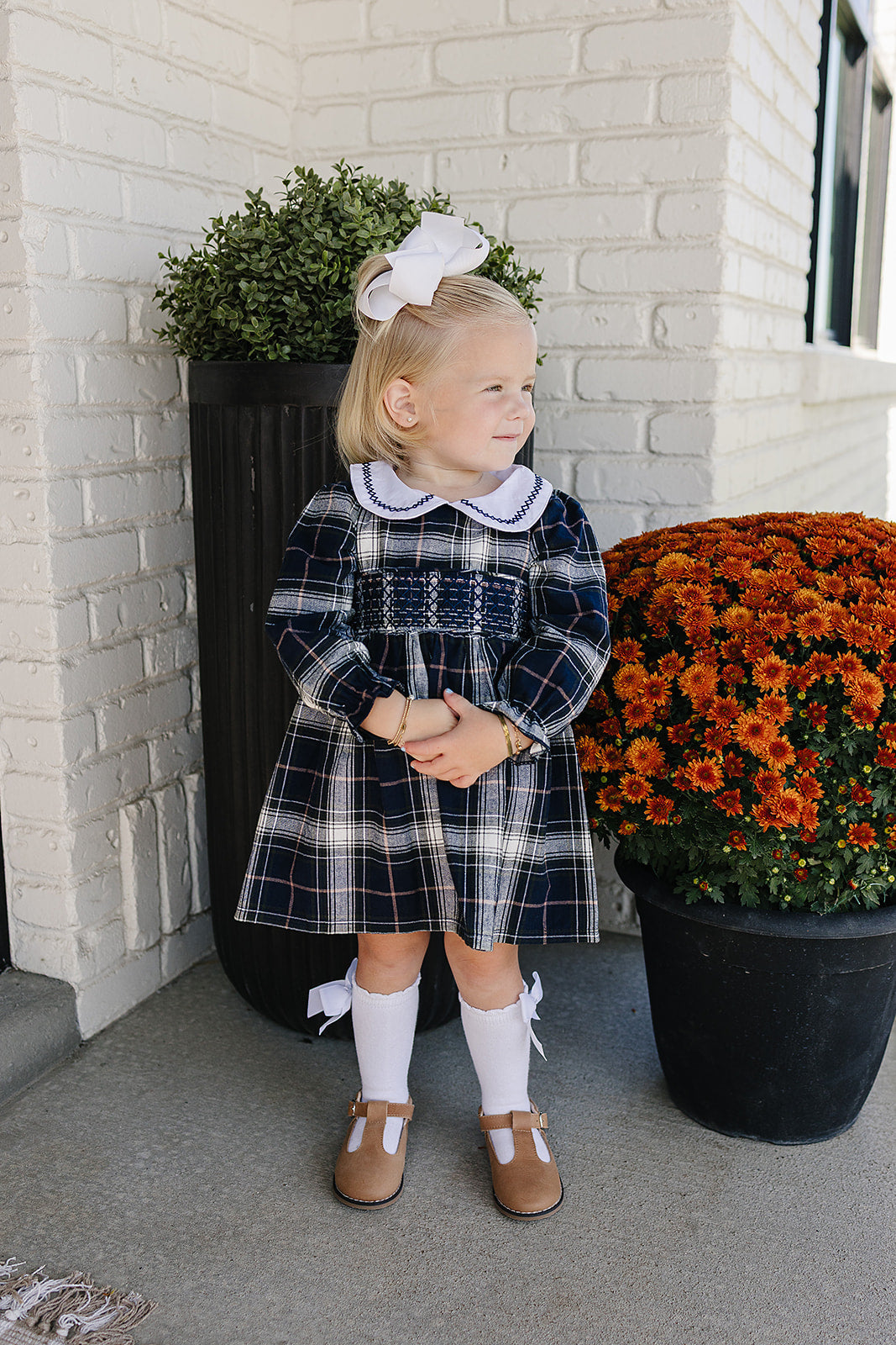 A girl wearing a Navy plaid smocked dress with Peter Pan collar, standing next to vibrant orange flowers.