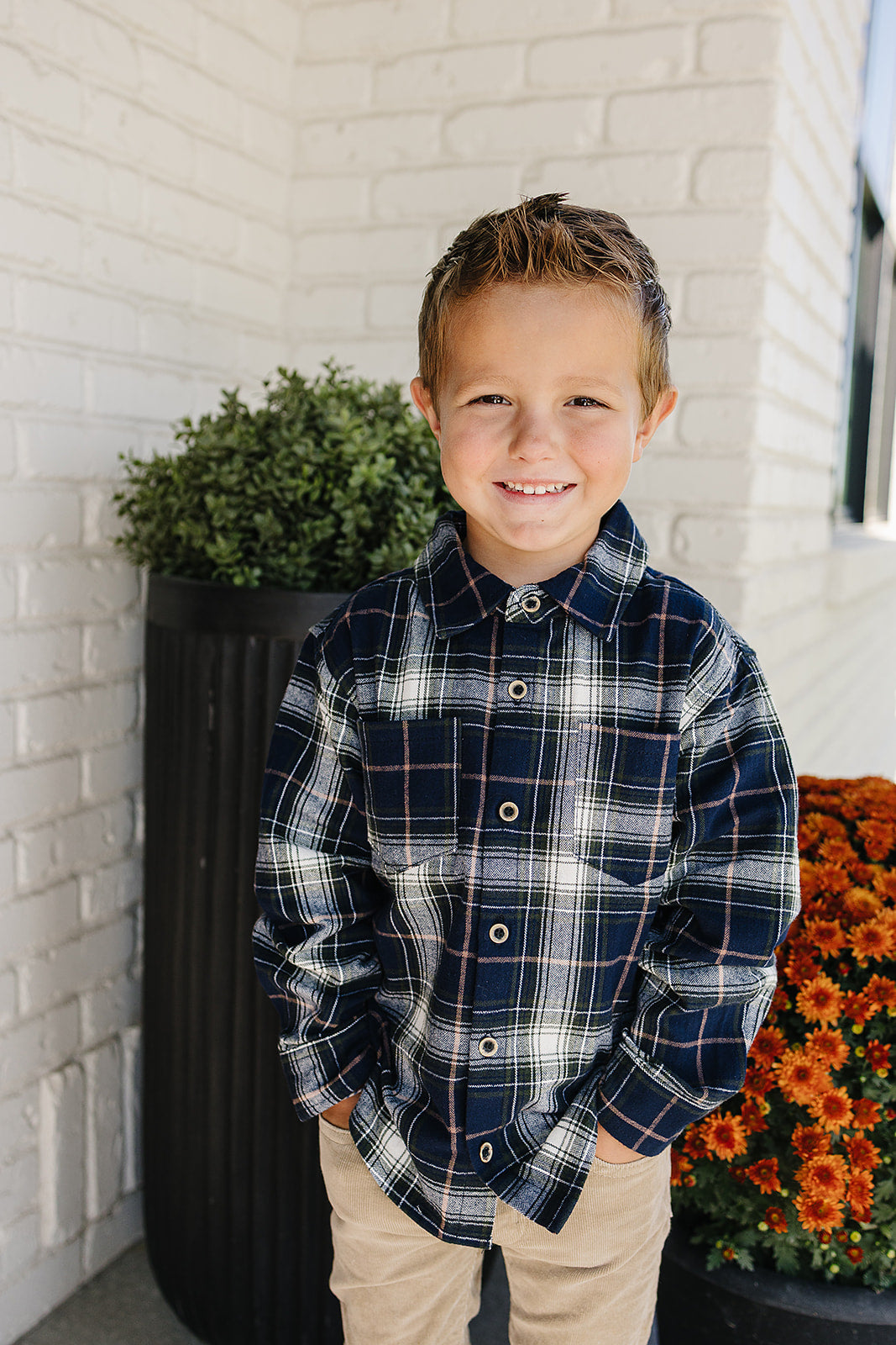 Smiling boy wearing a navy plaid button-up shirt for kids, standing in front of flowers and a brick wall.