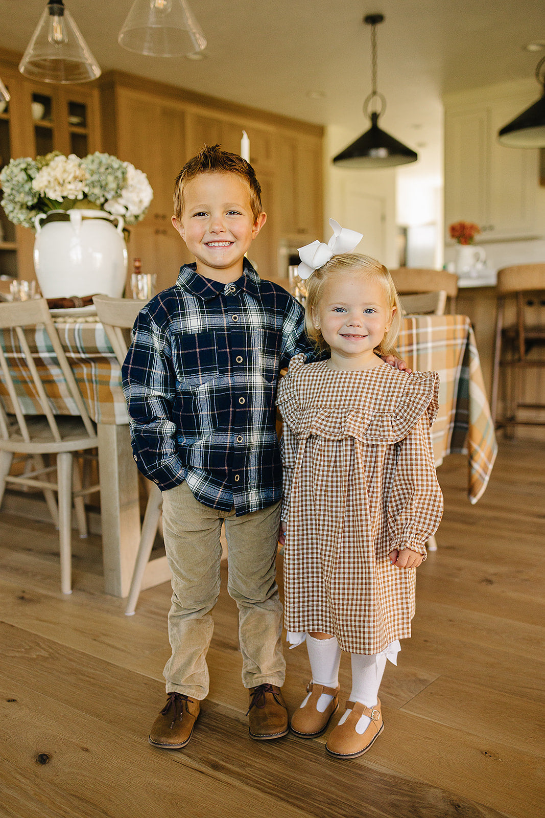 Two kids smiling in a cozy kitchen, one in a navy plaid button-up shirt and the other in a stylish dress.