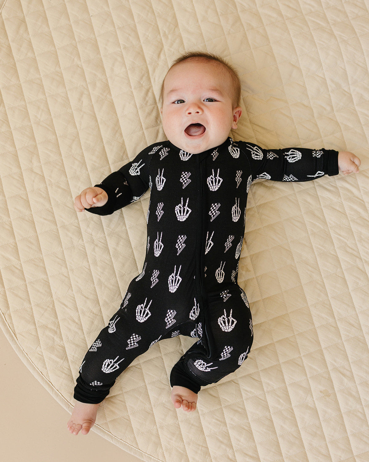 Baby wearing Bamboo Zip Romper Skelly Vibes with playful skeleton graphics on a quilted surface.