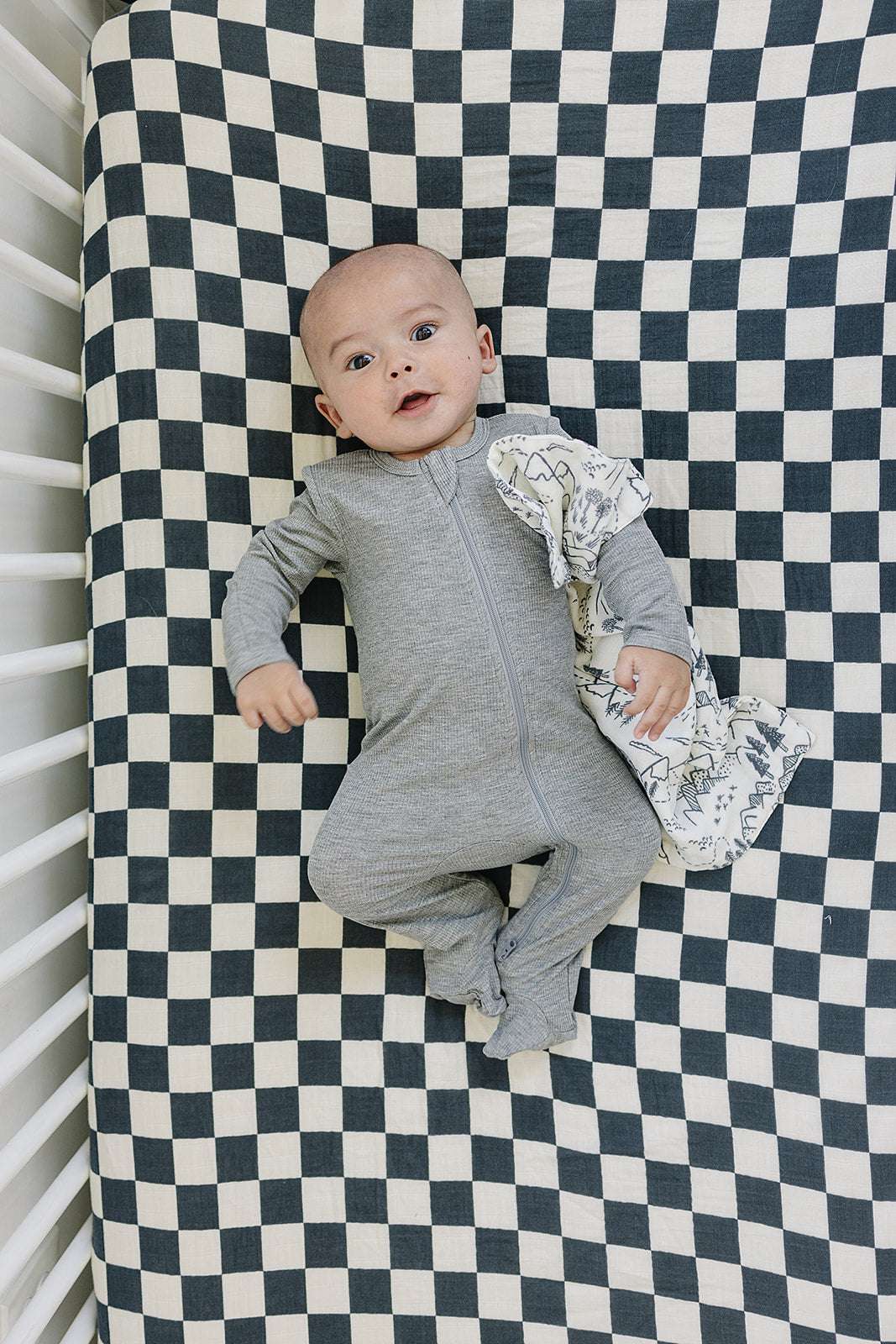 Baby lying on a charcoal checkered Muslin Crib Sheet, smiling while holding a cozy blanket.