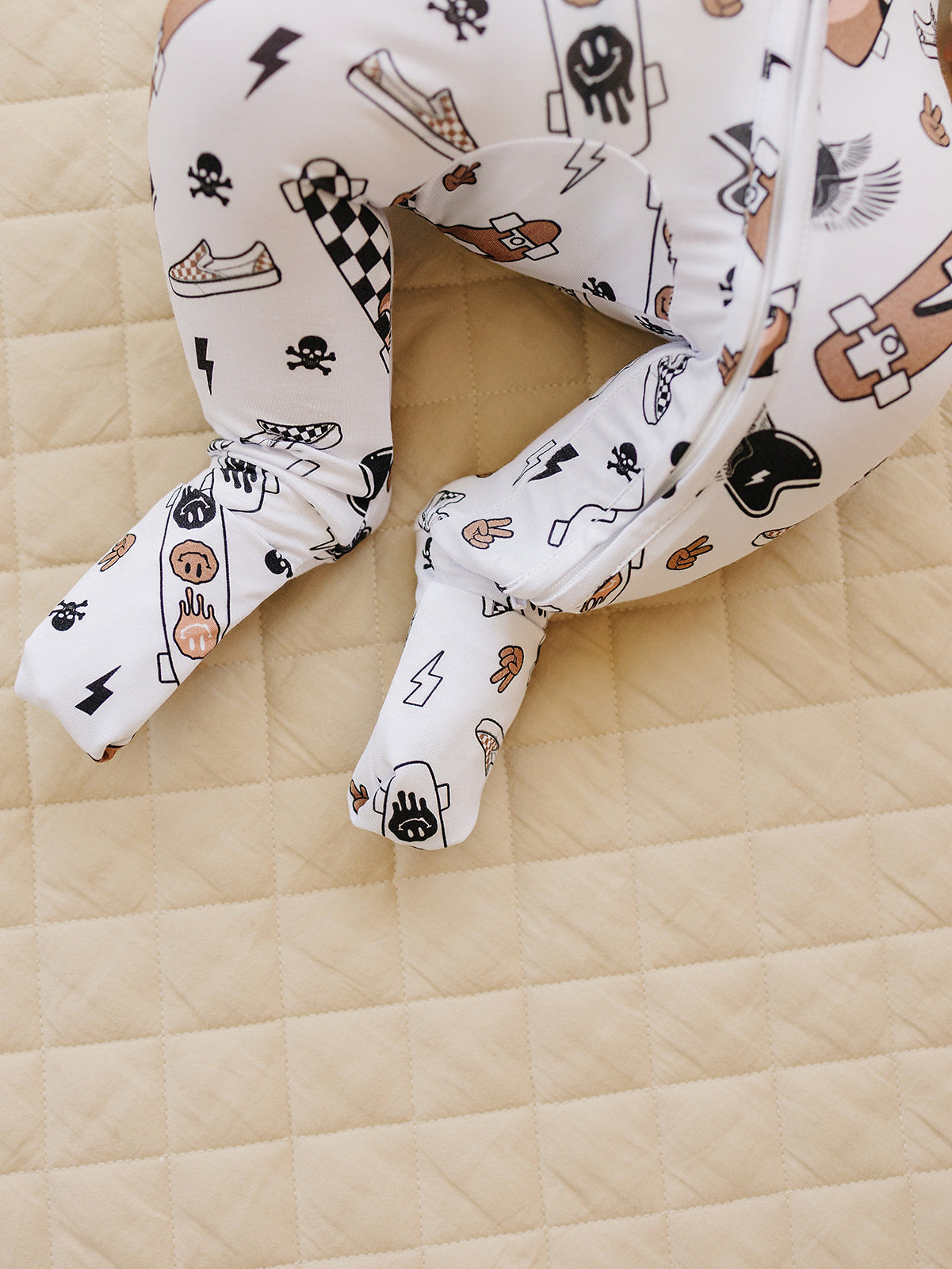 Close-up of Bamboo Zip Romper Sk8er with skateboard and graffiti designs on a beige quilted surface.