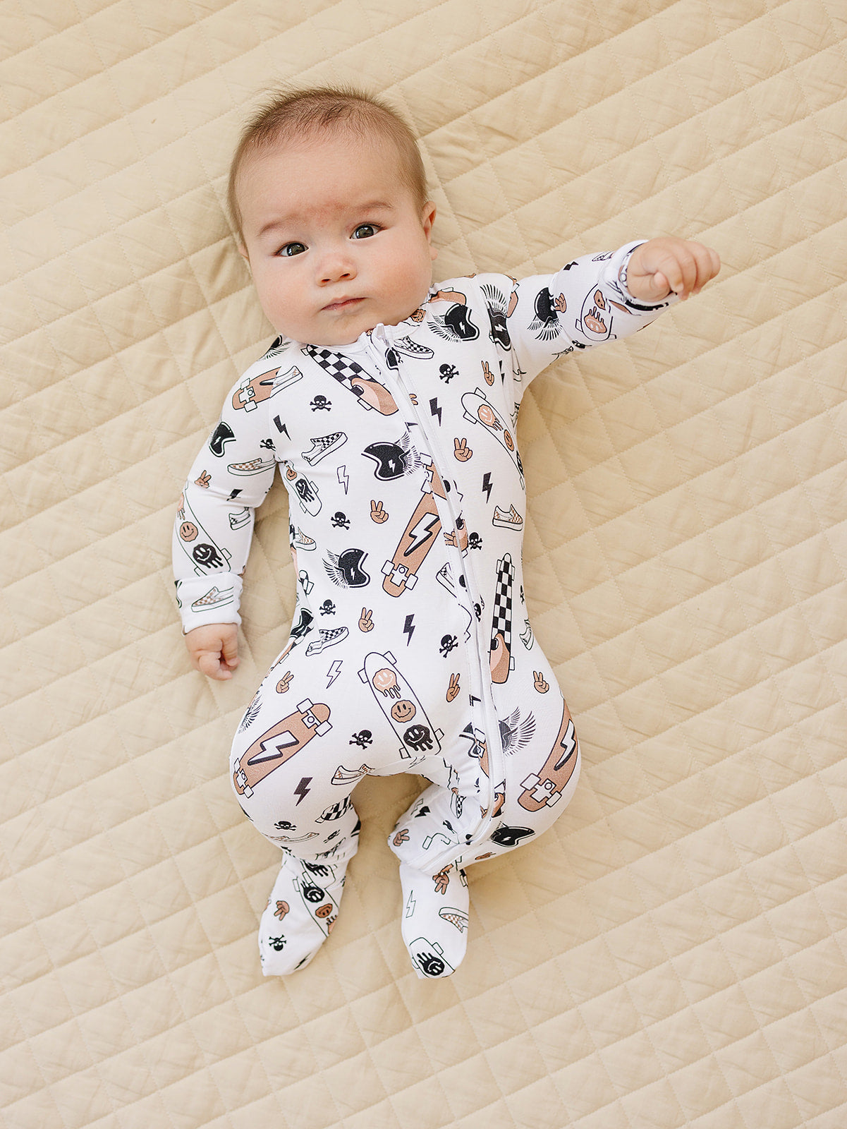 Baby wearing Bamboo Zip Romper Sk8er with skateboards and graffiti prints, laying on a quilted surface.