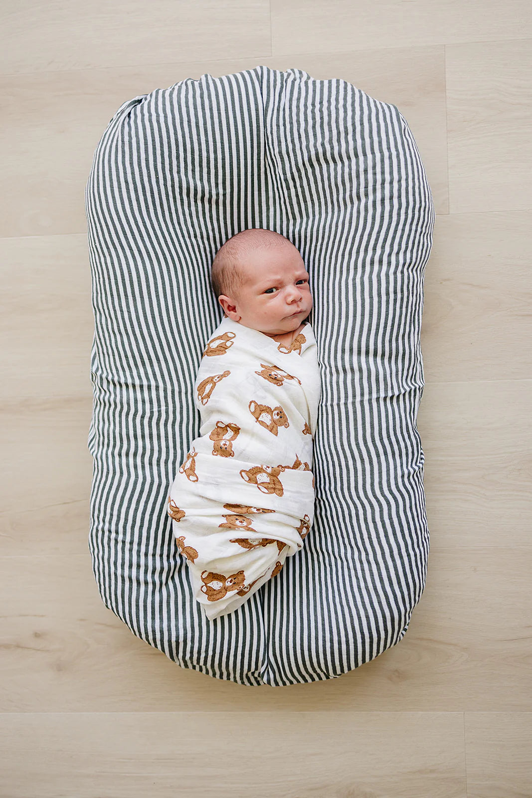 Baby swaddled in teddy bear muslin swaddle blanket, resting on a striped baby lounger.