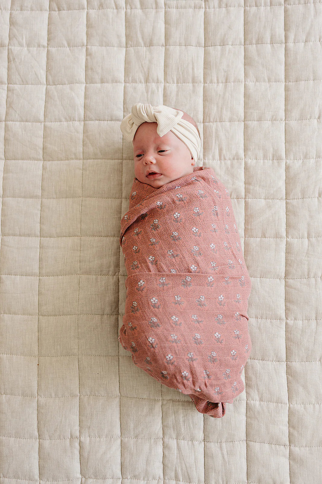 Baby wrapped in vintage pink floral muslin swaddle blanket with a headband on a neutral quilted background.