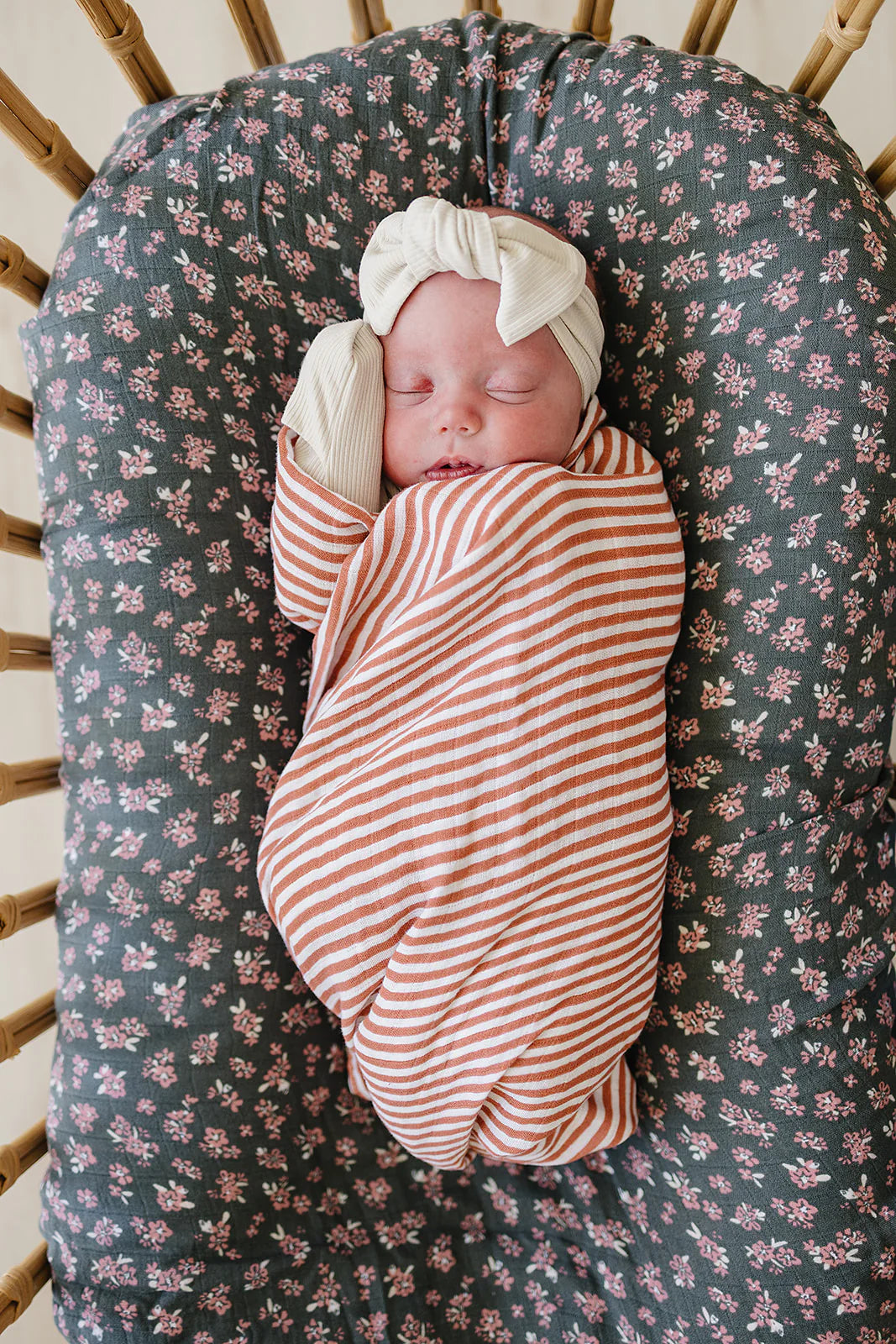 Newborn wrapped in a pink stripes muslin swaddle blanket, resting peacefully in a floral crib.