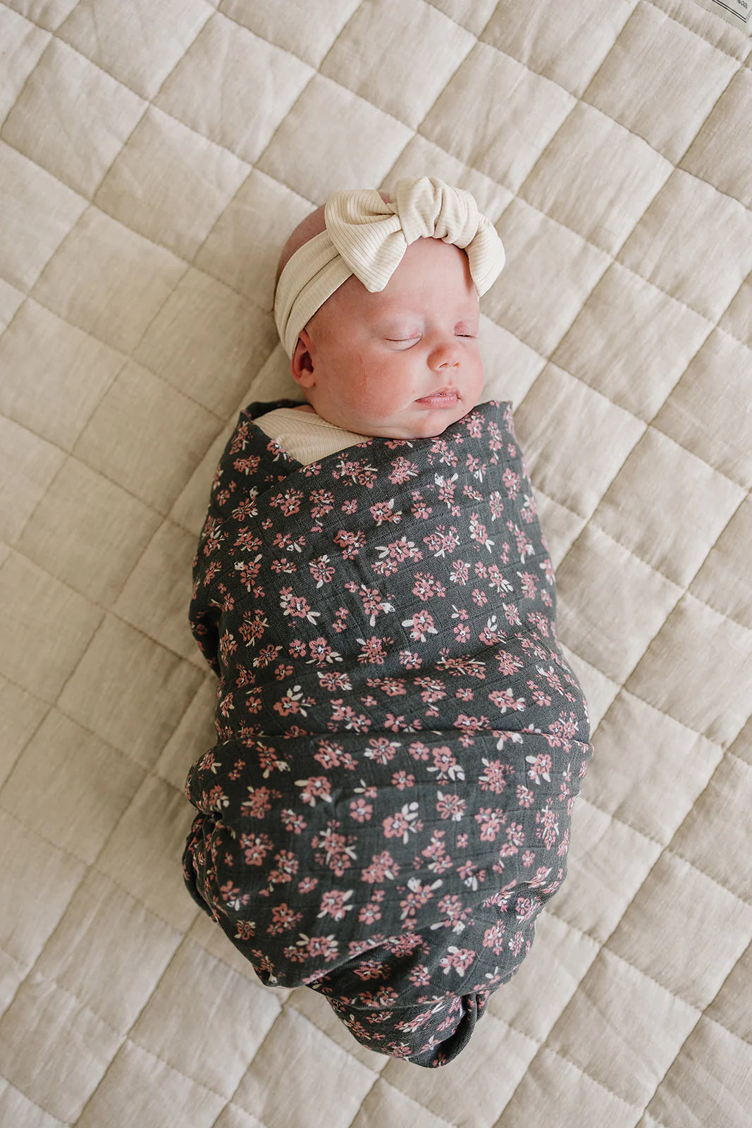 Baby peacefully swaddled in olive bloom muslin swaddle blanket on a quilted surface.