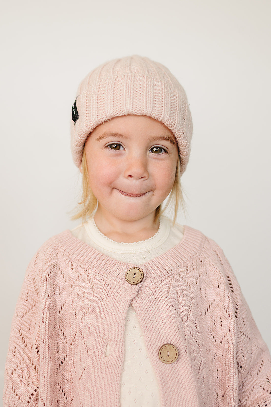 Child wearing a light pink chunky knit baby beanie, smiling and dressed in a matching pink cardigan.