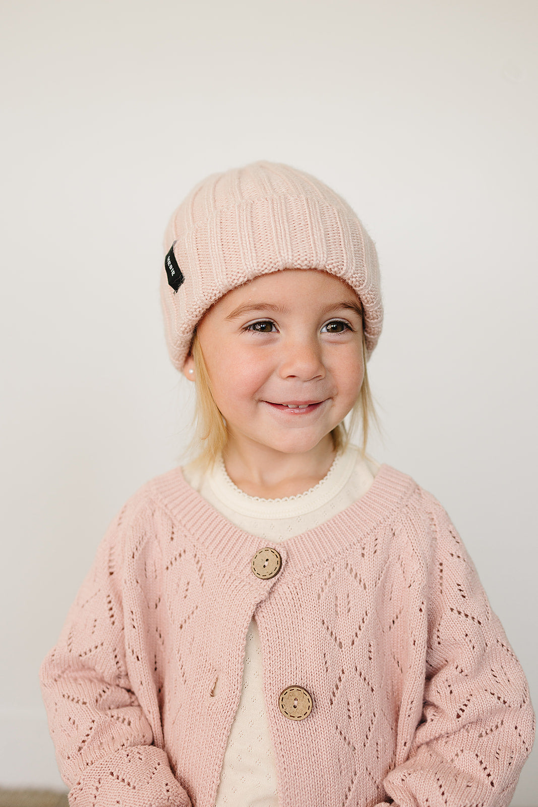 Smiling toddler wearing a light pink baby beanie and a pink knitted cardigan.