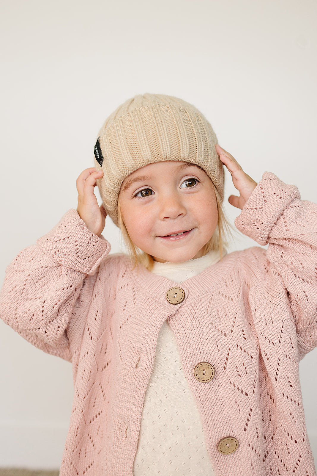 Smiling toddler wearing a tan Baby Beanie and a pink knitted cardigan, showcasing cozy winter attire.