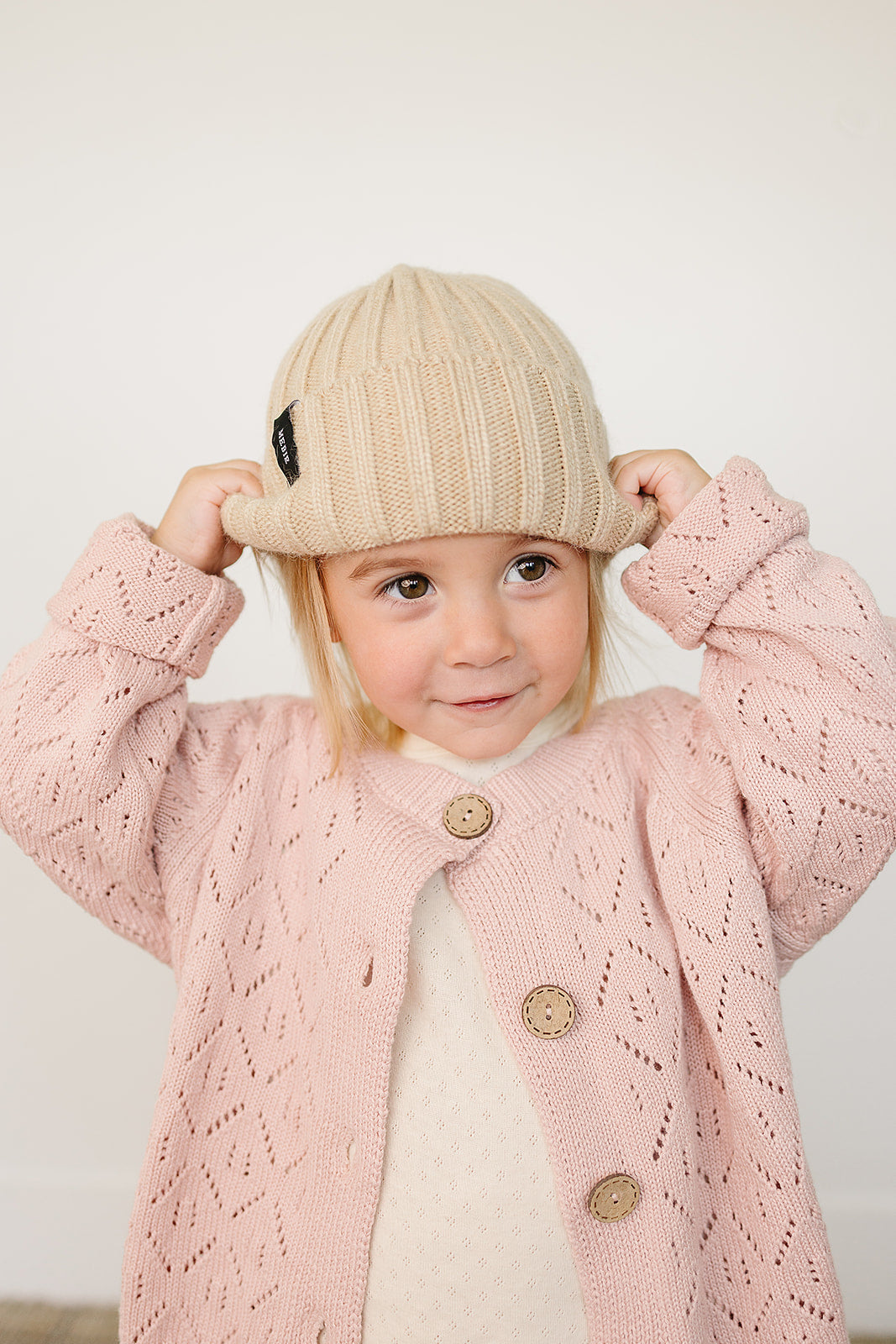 Baby wearing a tan chunky knit baby beanie, smiling while adjusting the cozy hat with a pink cardigan.