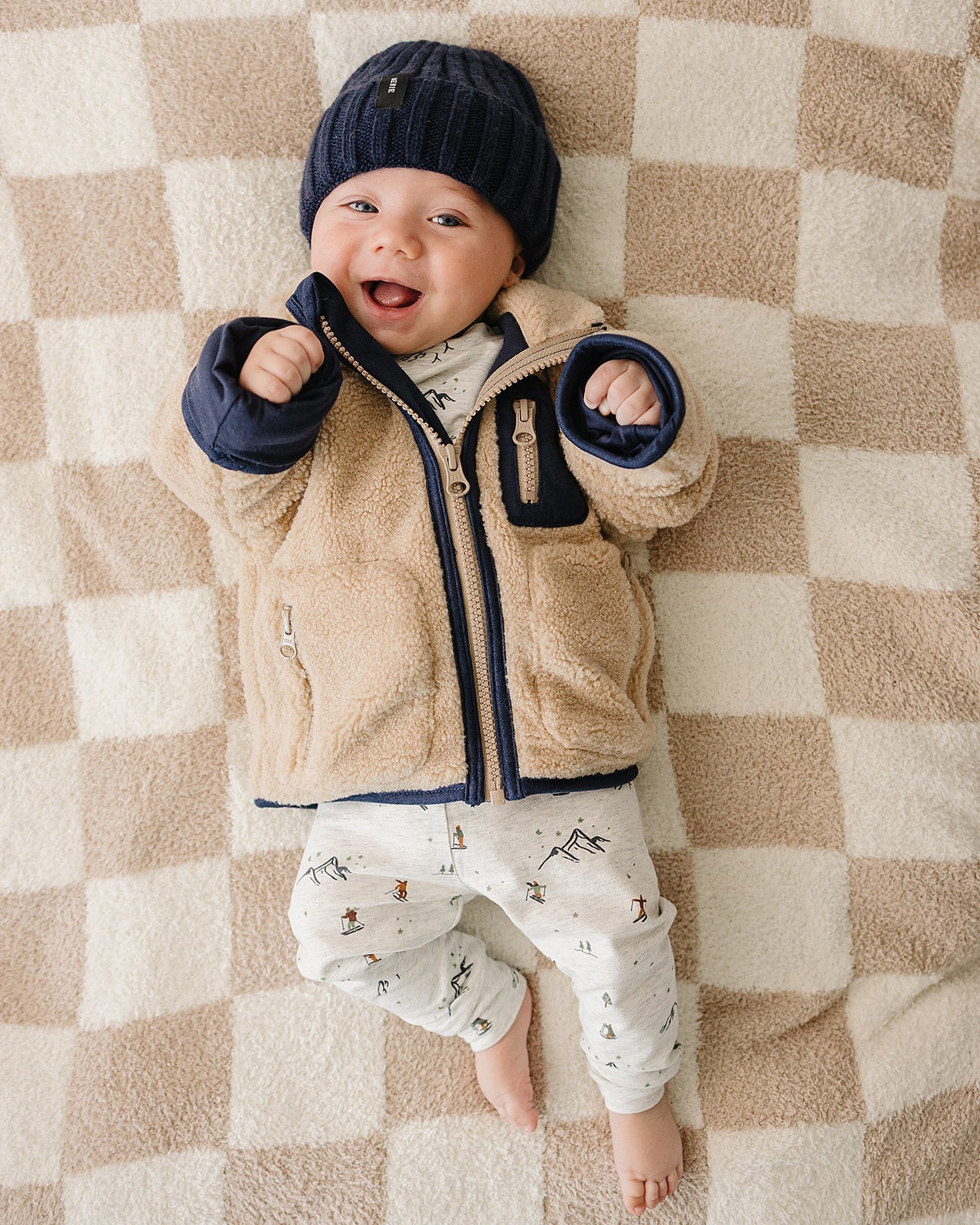 Happy baby in a cozy chunky knit baby beanie and winter jacket on a blanket.