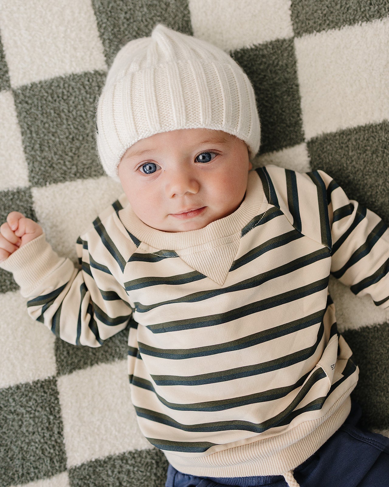 Baby wearing a chunky knit baby beanie in white, cozy outfit with stripes, on a checkered blanket.