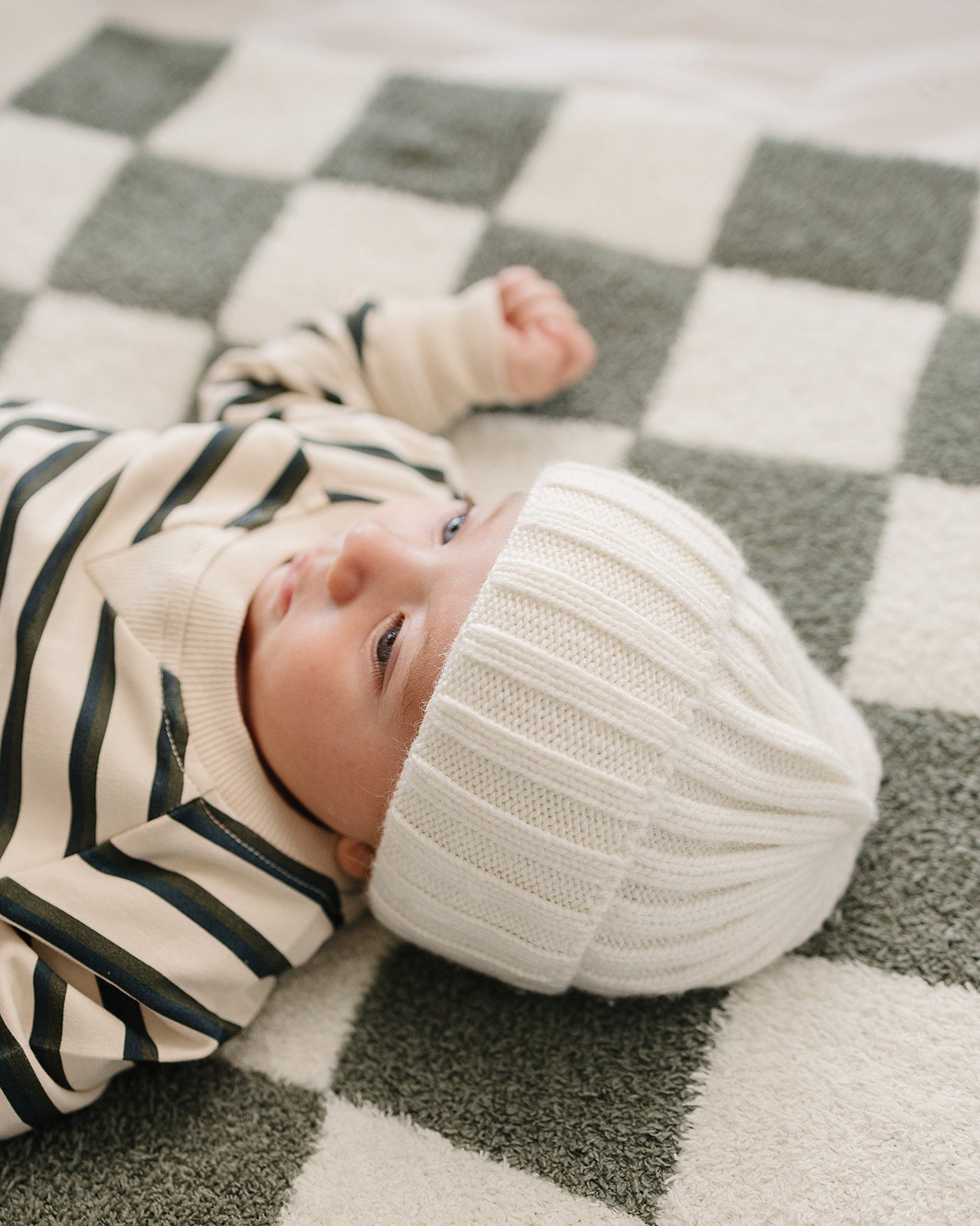 Baby wearing a chunky knit baby beanie in white, lying on a checkered blanket.