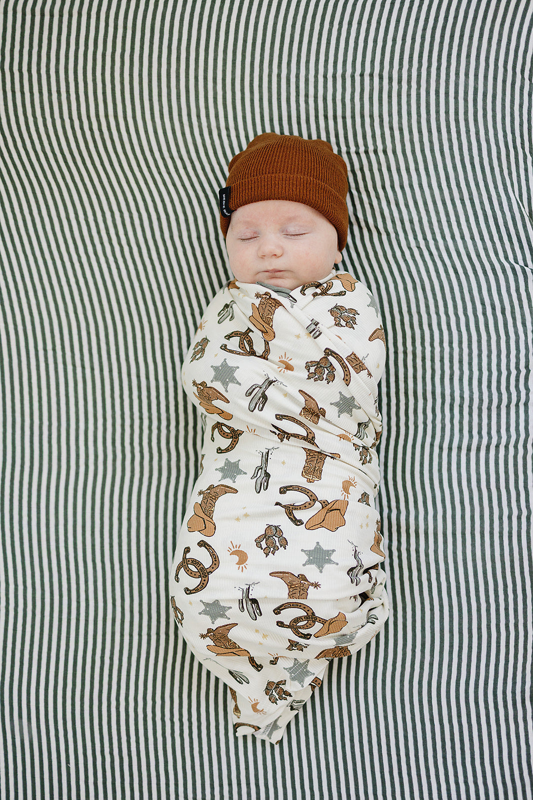 Baby wrapped in Bamboo Stretch Swaddle Outlaw Cowboy Ribbed with a brown hat, laying on a striped blanket.