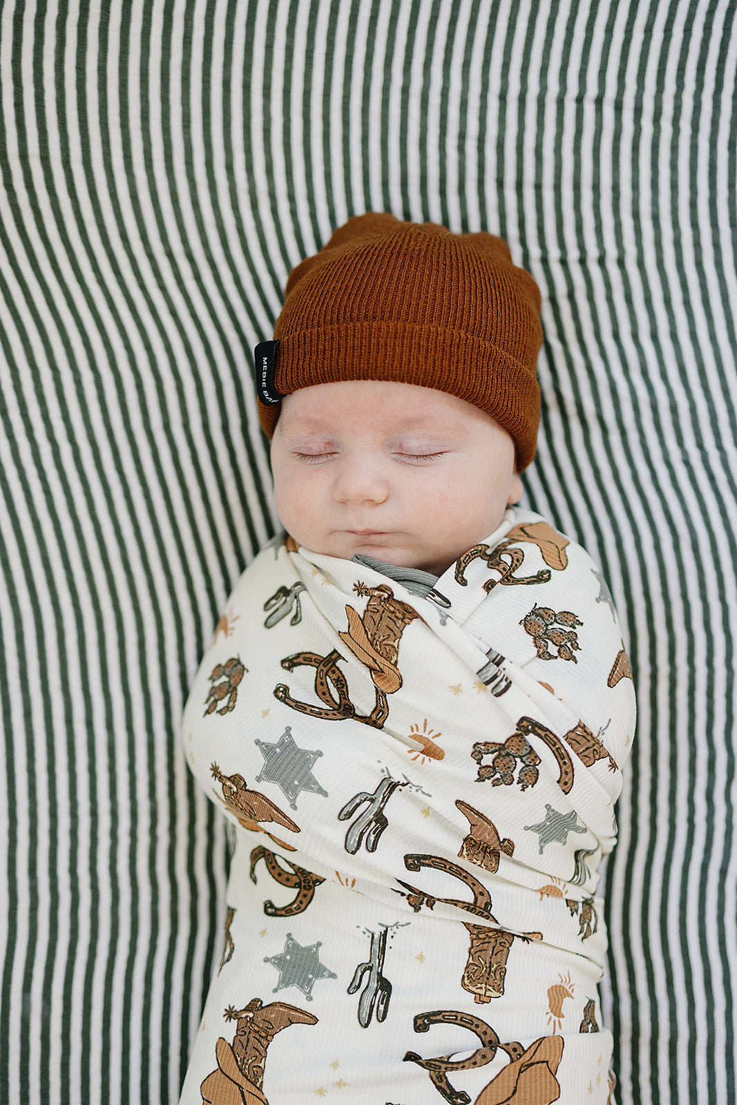 Baby wrapped in Bamboo Stretch Swaddle Outlaw Cowboy Ribbed with a brown hat, sleeping peacefully on striped bedding.
