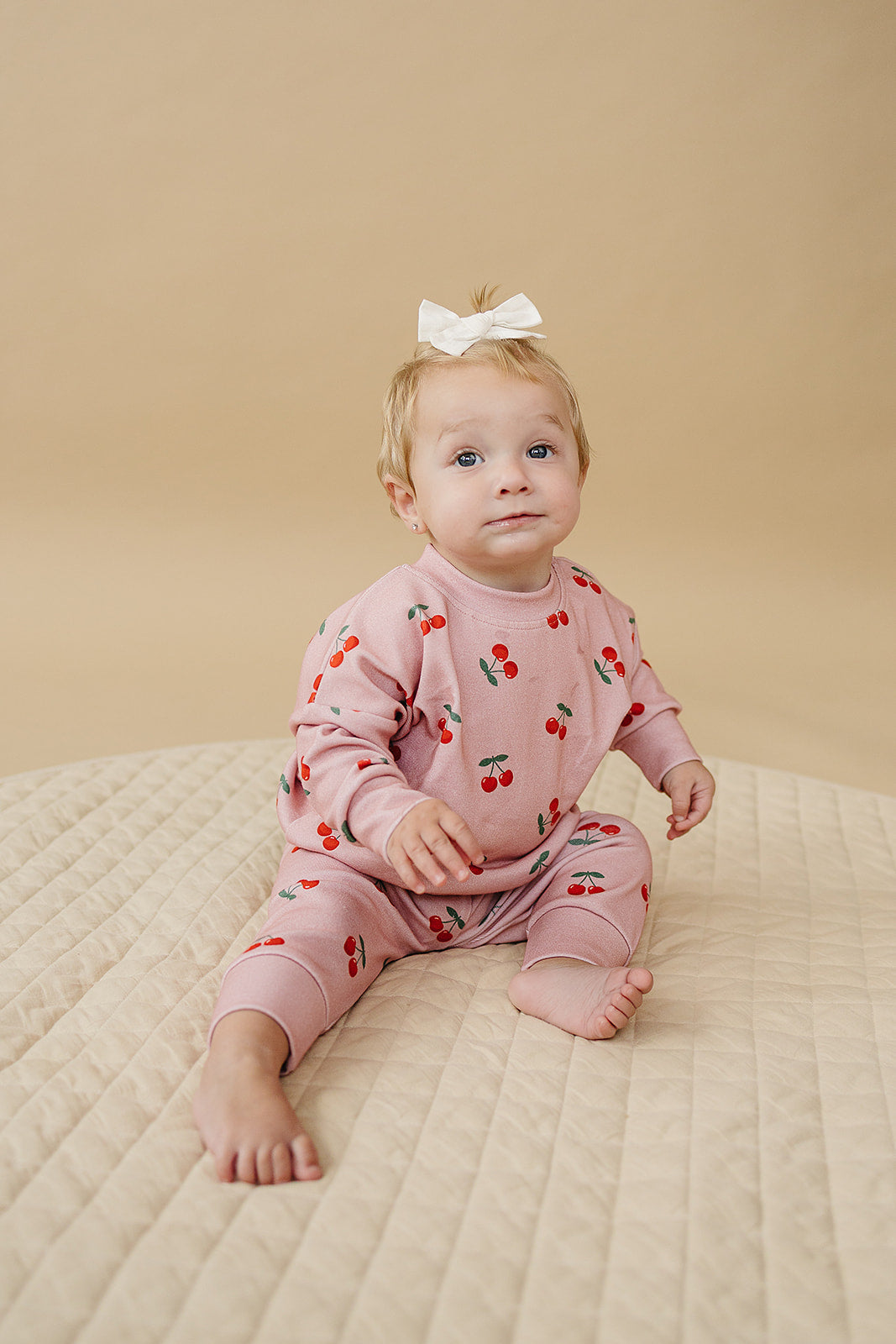 Blush Cherry Kids Lounge Set on a baby girl sitting on a quilted surface, wearing a cherry print outfit and a bow.