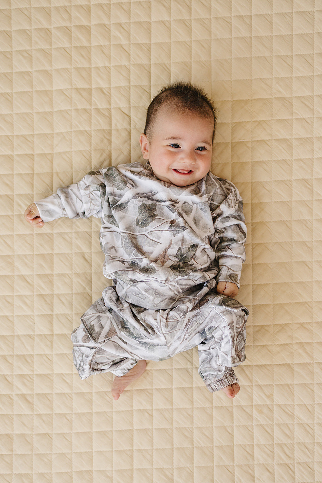 Camo kids lounge set featuring a baby smiling in camo pullover hoodie and cargo jogger pants on a quilted blanket.
