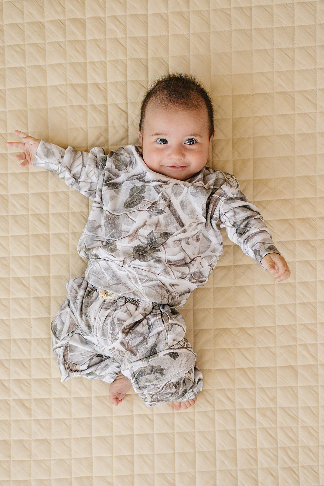 Camo kids lounge set featuring a baby wearing a pullover hoodie and cargo joggers on a quilted surface.