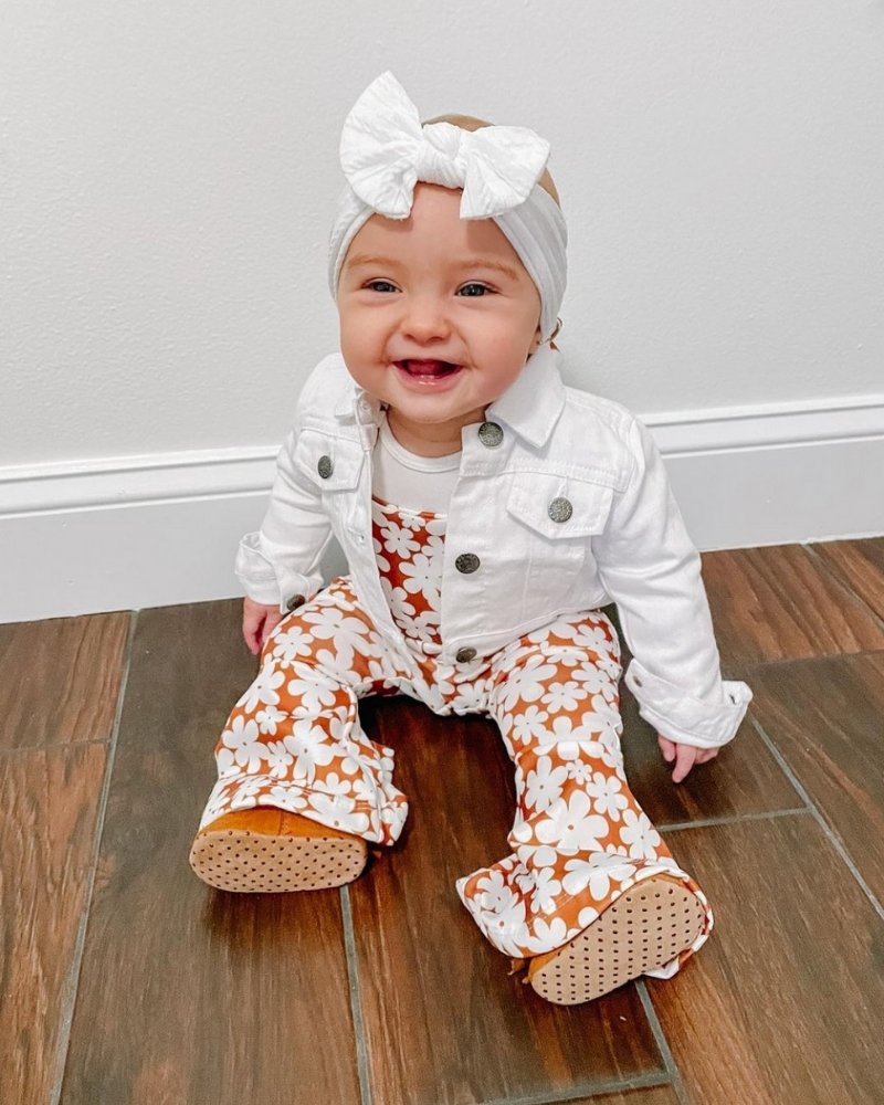 Baby wearing Andi pleated bell bottom jumpsuit in flower print, smiling with a white jacket and headband.