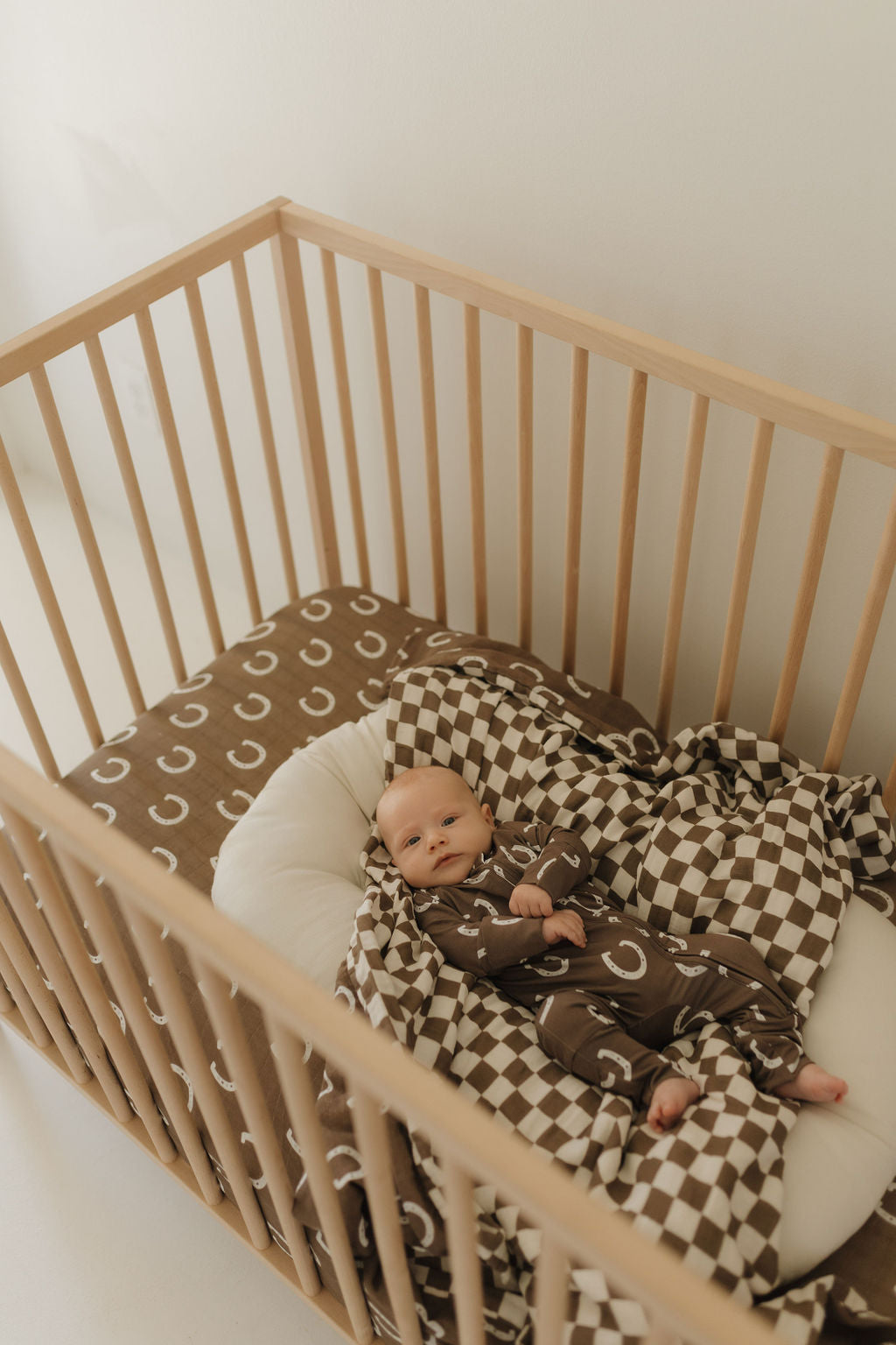 Infant in bamboo zip pajamas lying in a crib with brown patterned bedding and a cozy pillow.