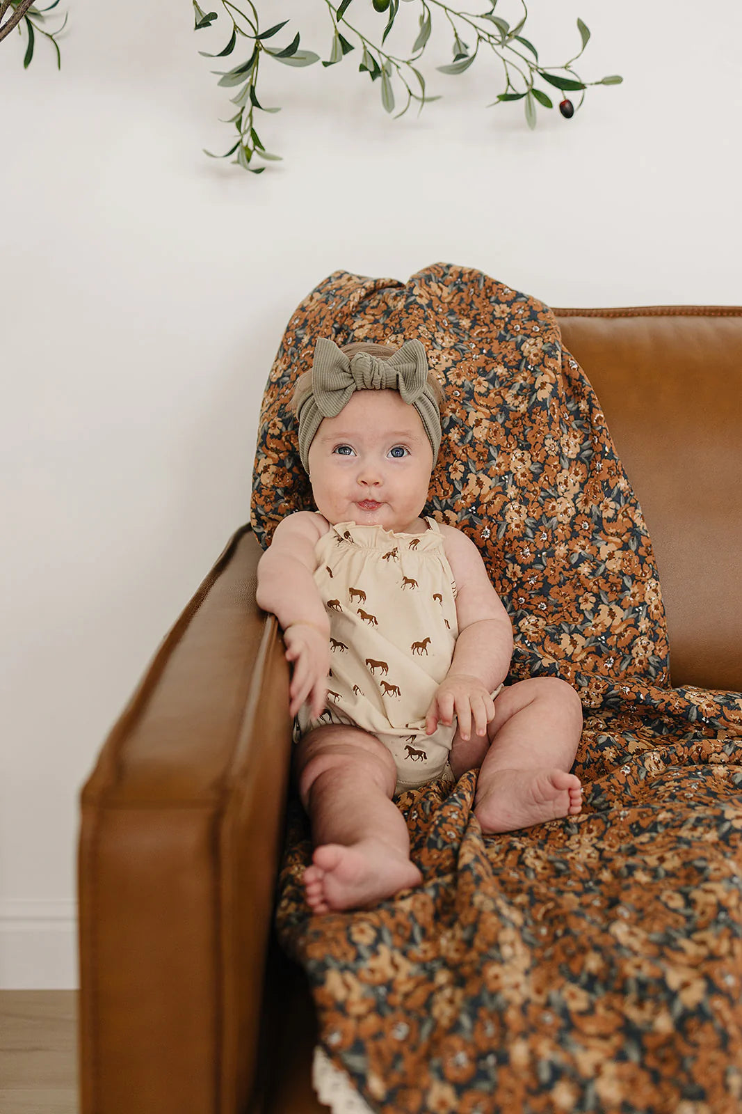 Baby girl in horse print bubble romper with a green headband on a cozy couch.