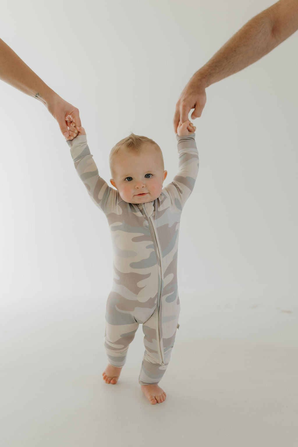 Baby wearing bamboo zip pajamas in neutral camo, held by parents, showcasing comfort and modern design.