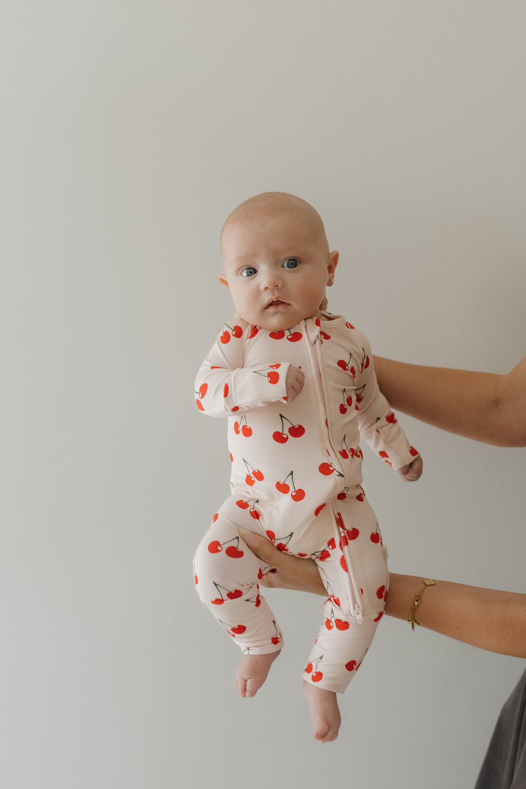 Baby wearing Cherry Orchard bamboo zip pajamas with red cherry print, held by an adult hand.