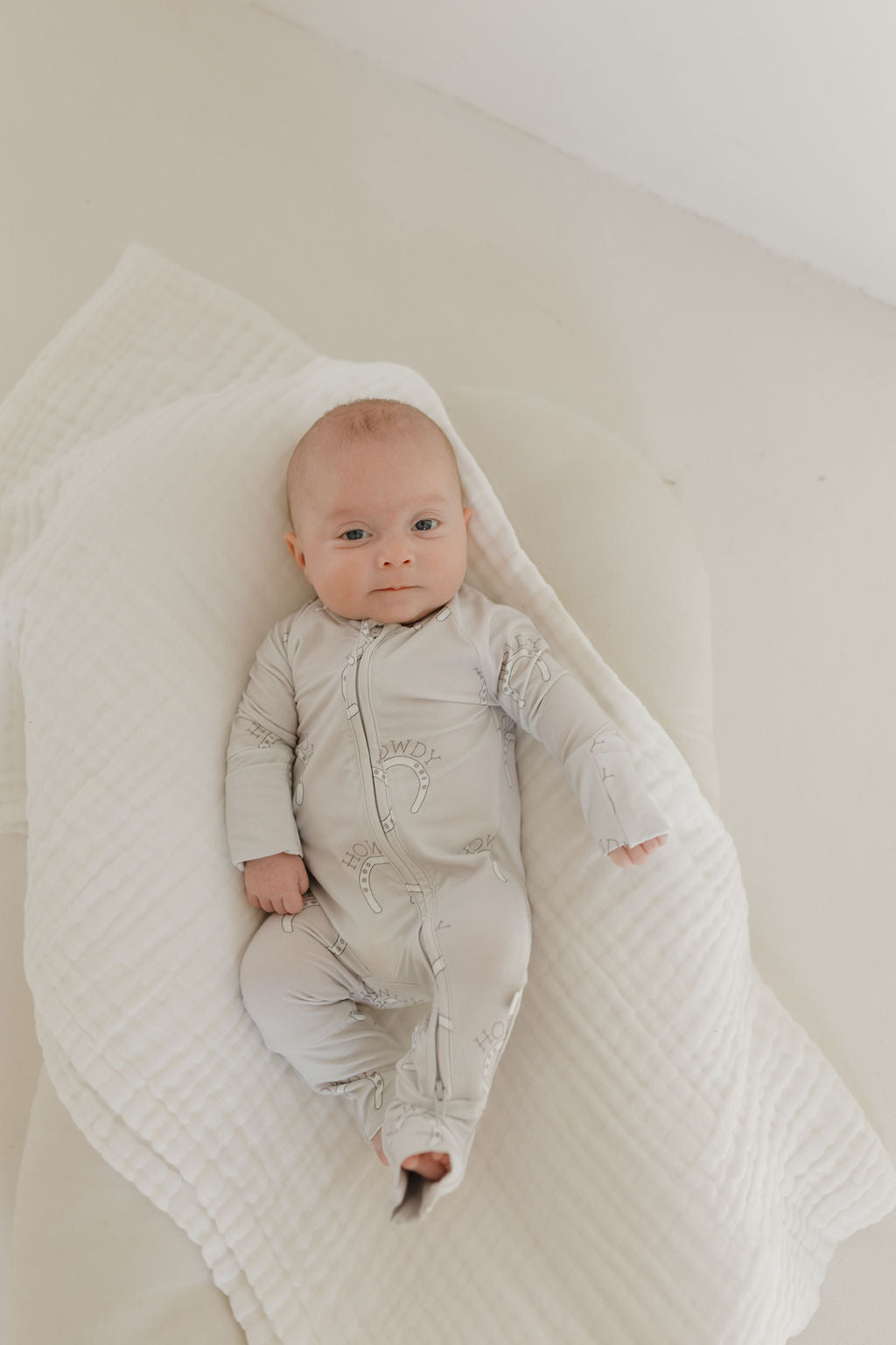 Infant wearing bamboo zip pajamas with a playful 'Howdy' design, resting on a soft blanket.