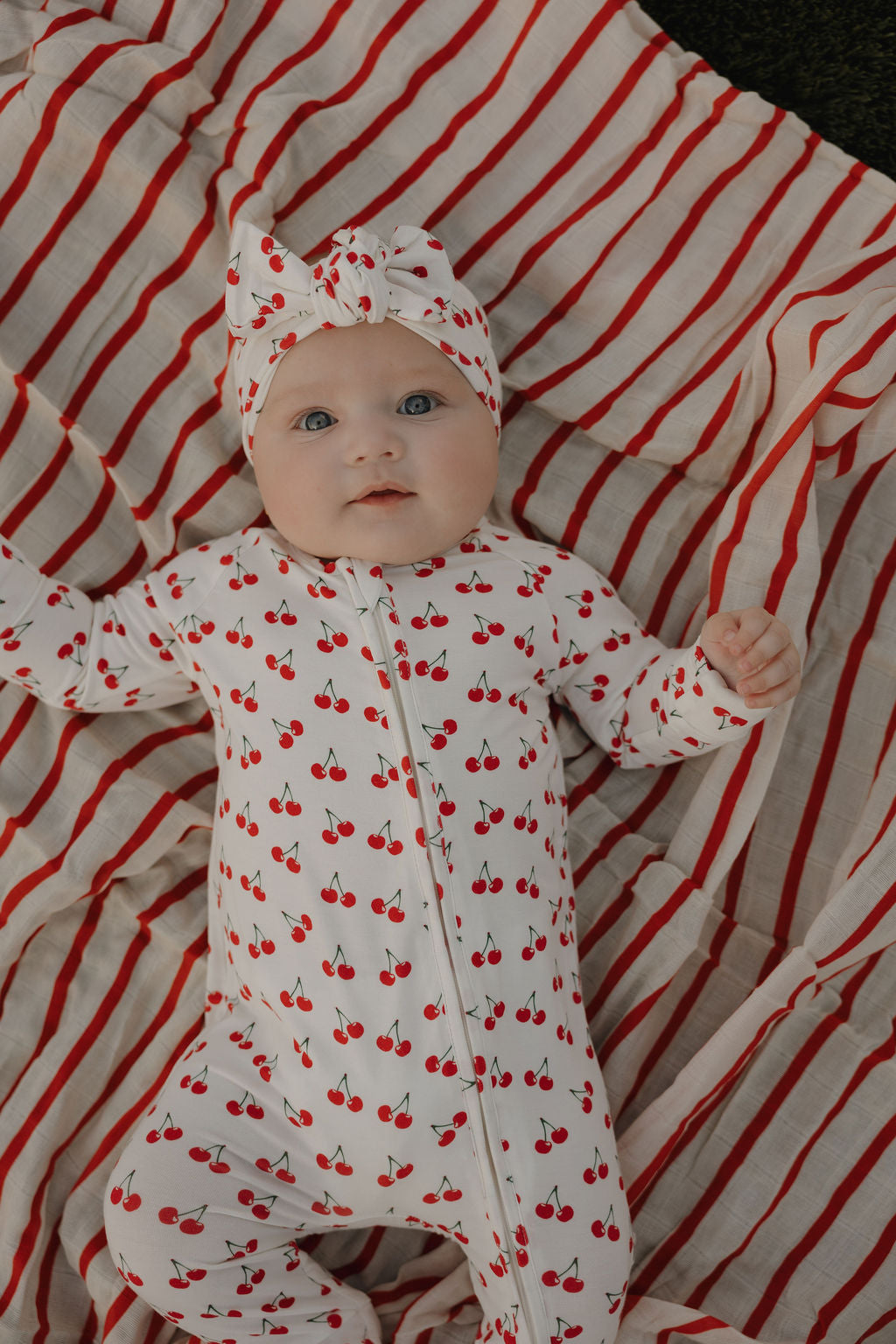 Baby in cherry-printed bamboo zip pajamas and matching headband on striped blanket.