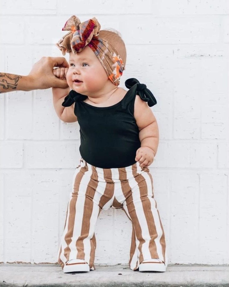 Cute baby wearing tan striped bell bottom pants and a black top with bows, styled with a colorful headband.