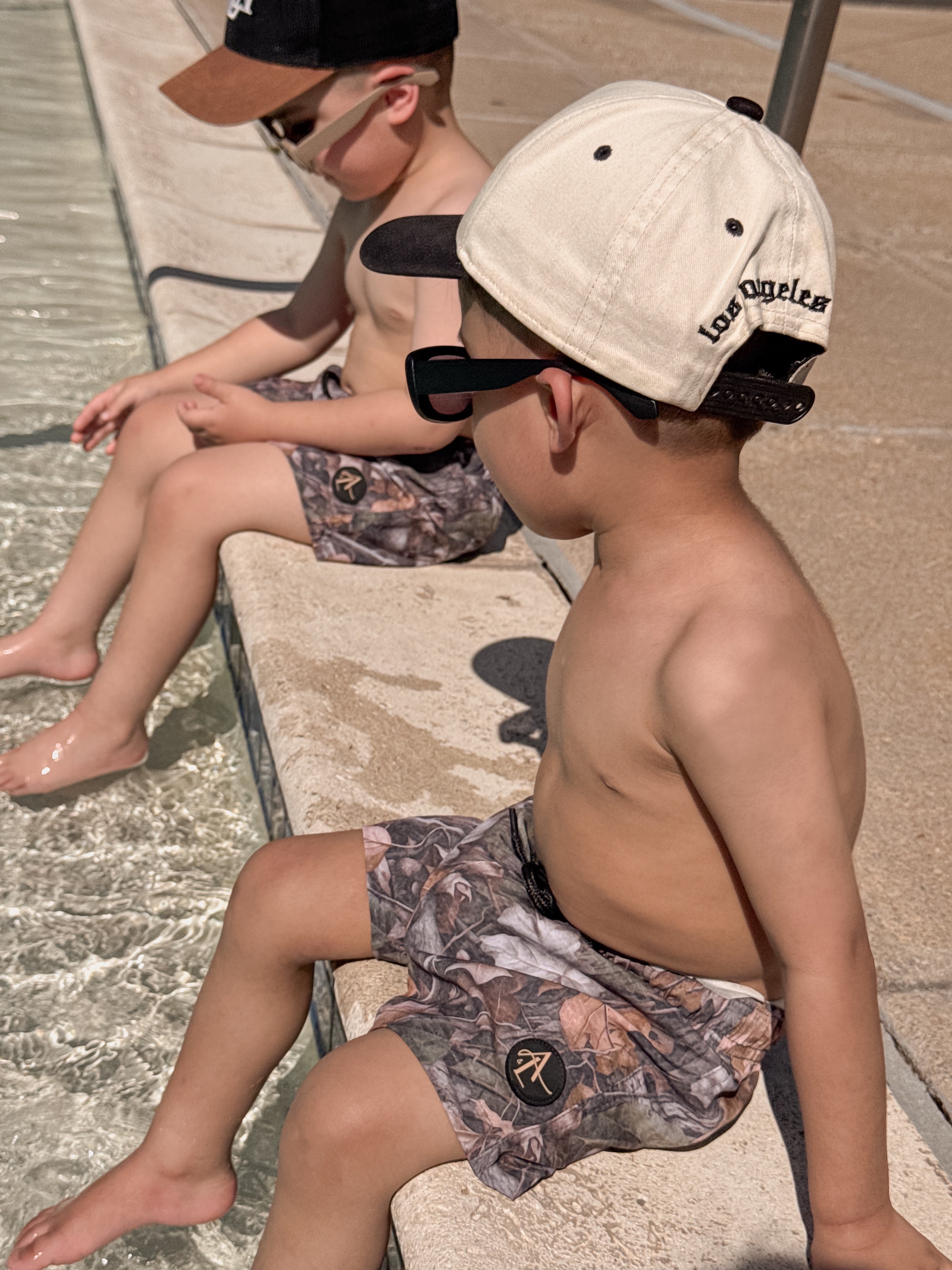 Two boys in camo swim shorts relaxing by the pool on a sunny day, enjoying water play and summer fun.