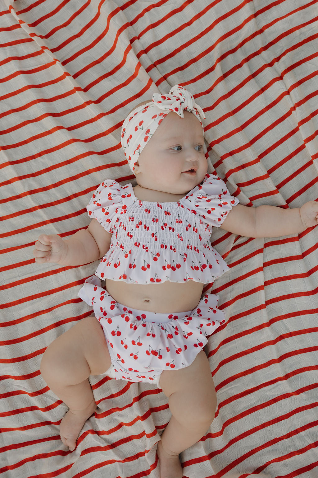 Cute baby in cherry print two-piece swimsuit with ruffle top and matching headband on a striped blanket.