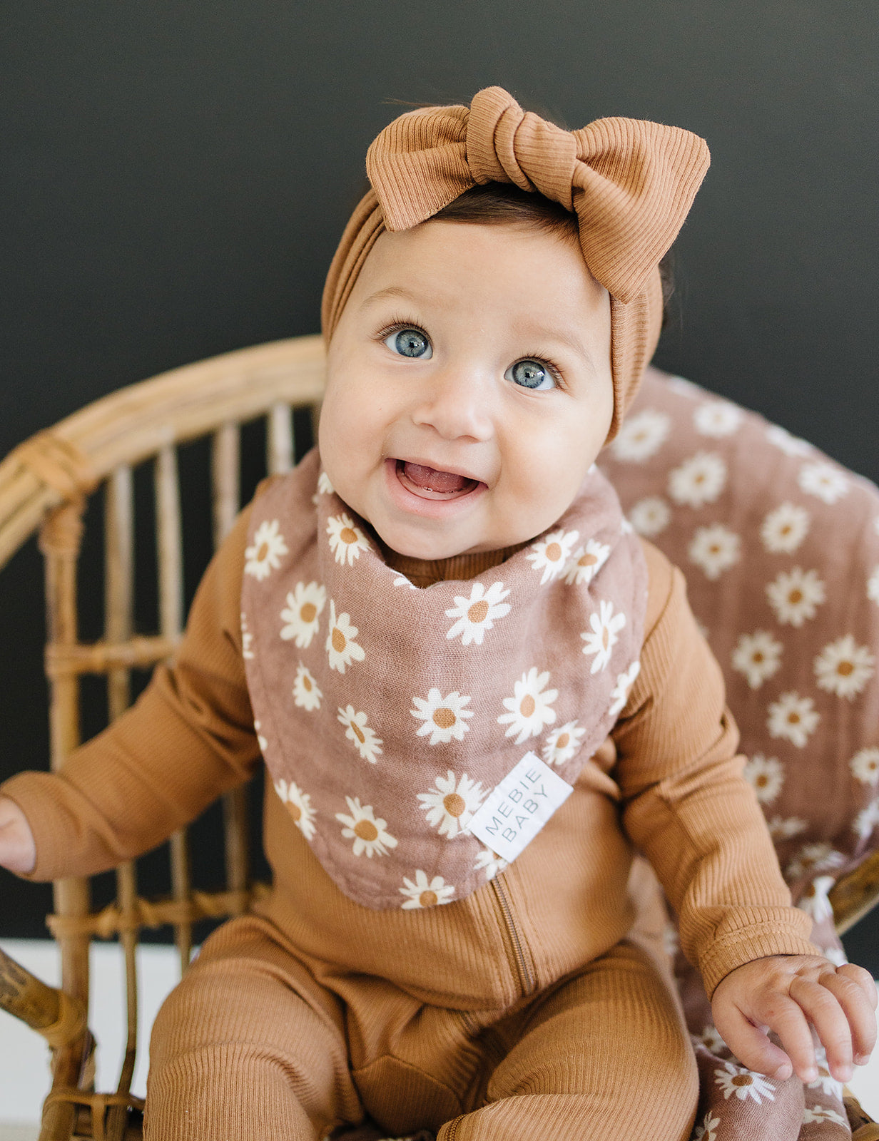 Happy baby wearing a daisy print muslin bib and matching headband, seated in a wicker chair.