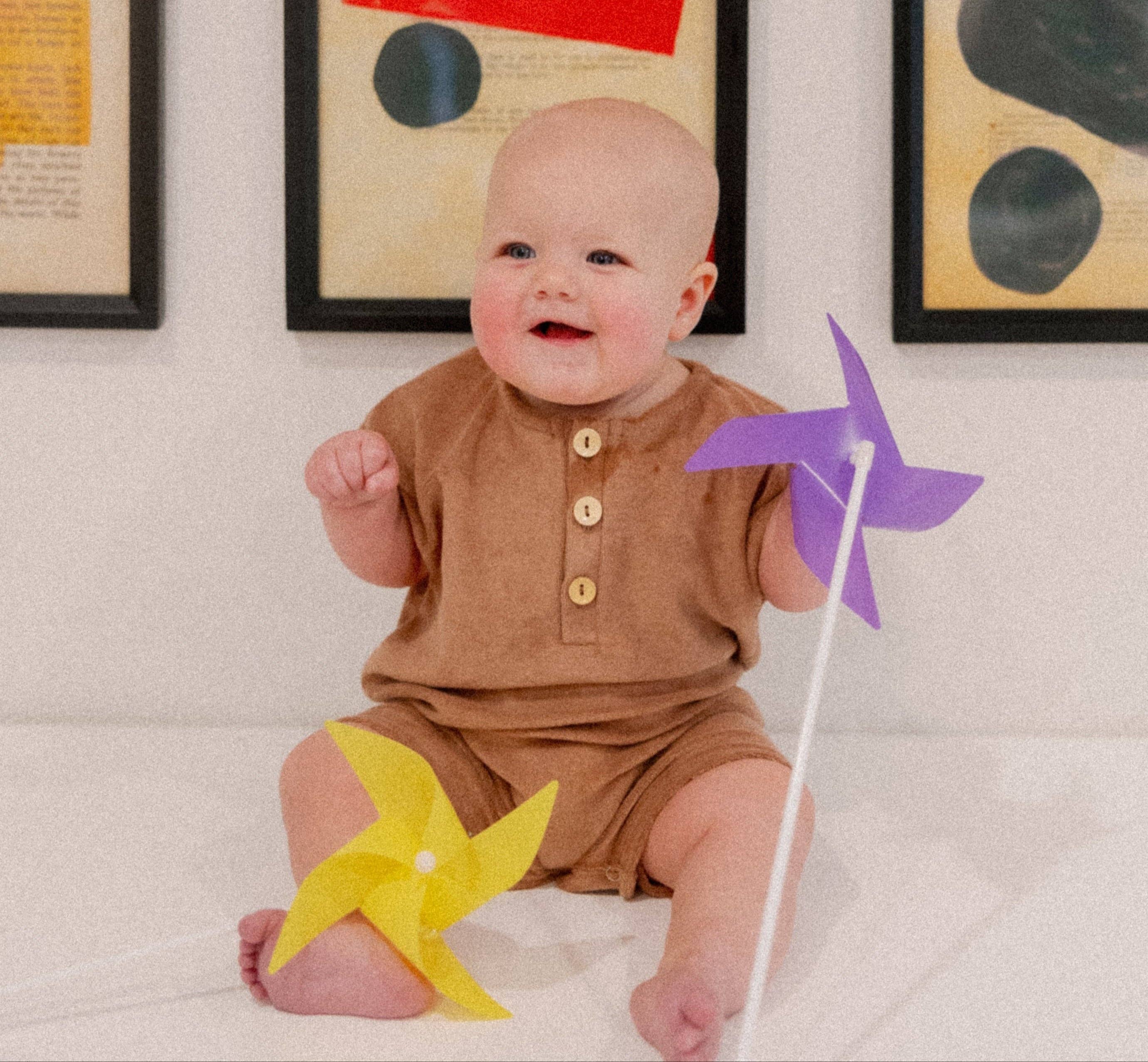 Cute baby wearing a brown Dillon Romper, holding colorful pinwheels, with artwork in the background.