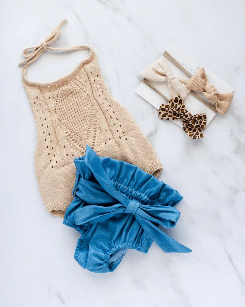 Drake crochet halter romper in vanilla cream with blue bow shorts and hair accessories on marble background.