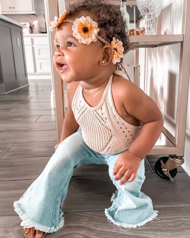 Cute baby girl in crochet halter romper and flared jeans, smiling with floral headband in a bright kitchen setting.