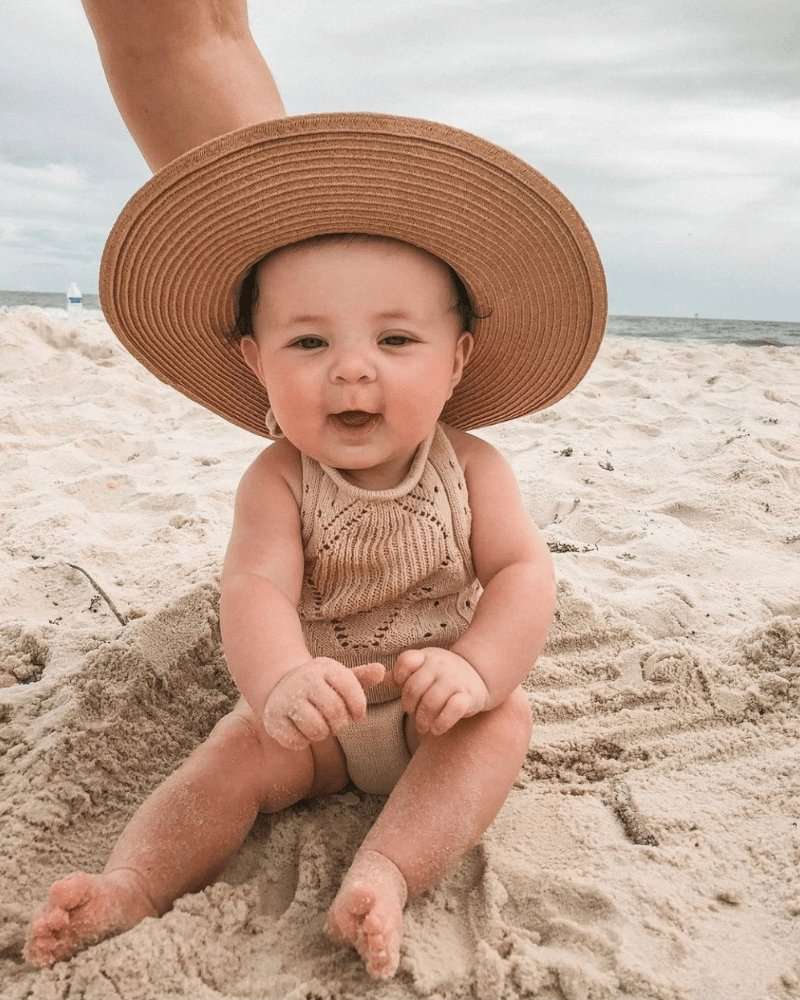 Adorable baby wearing a crochet swimsuit and sun hat sitting on the beach, enjoying a sunny day.