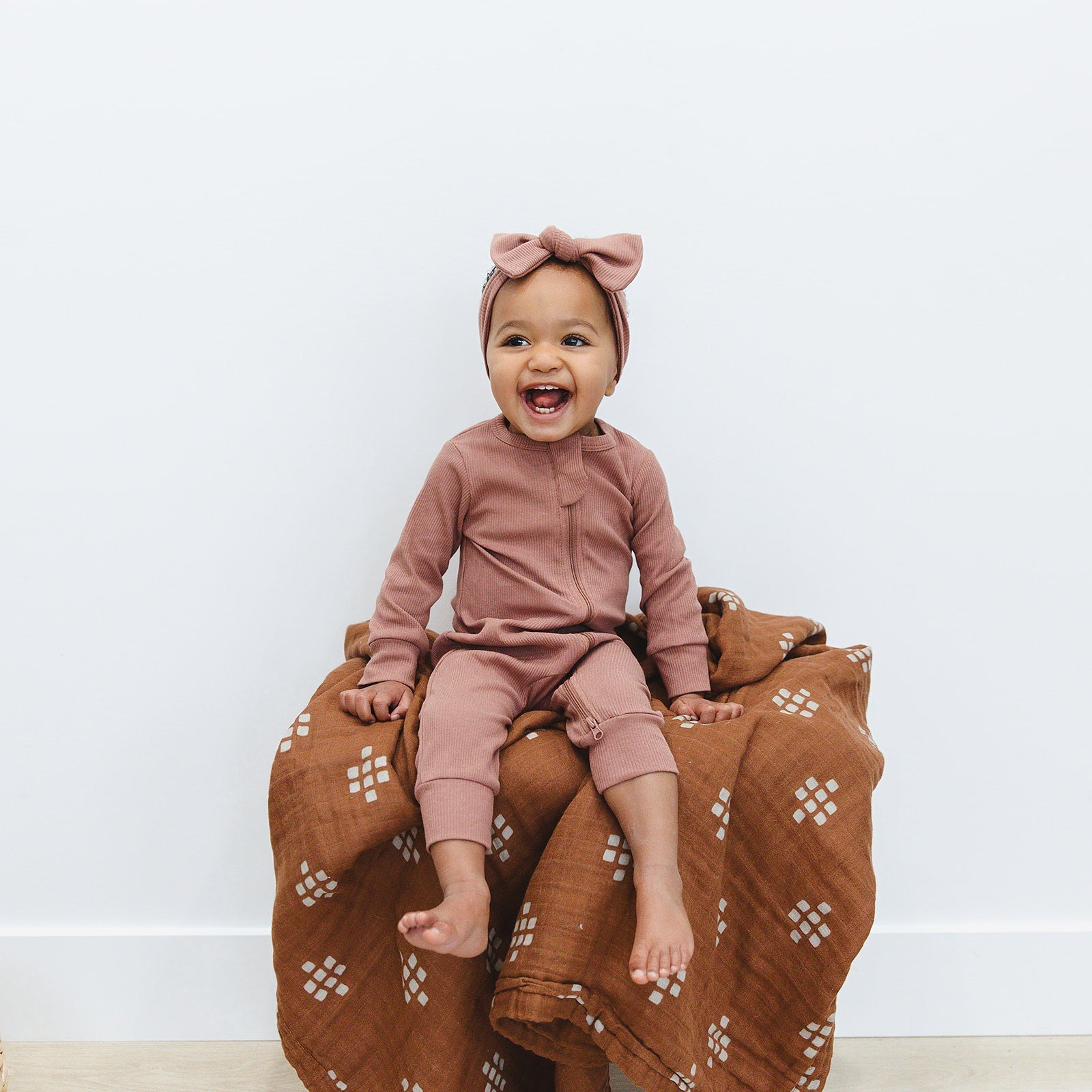 Smiling baby in dusty rose organic cotton ribbed head wrap and matching outfit, sitting on a blanket.
